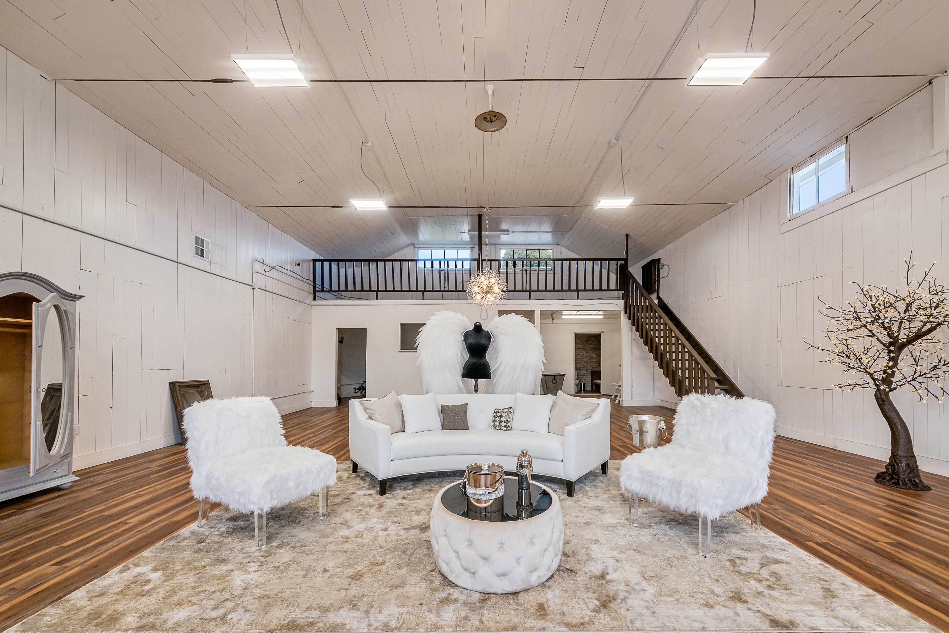 The spacious room features a white sofa facing a circular coffee table, surrounded by two plush chairs and a small decorative tree, with a staircase and large windows in the background.