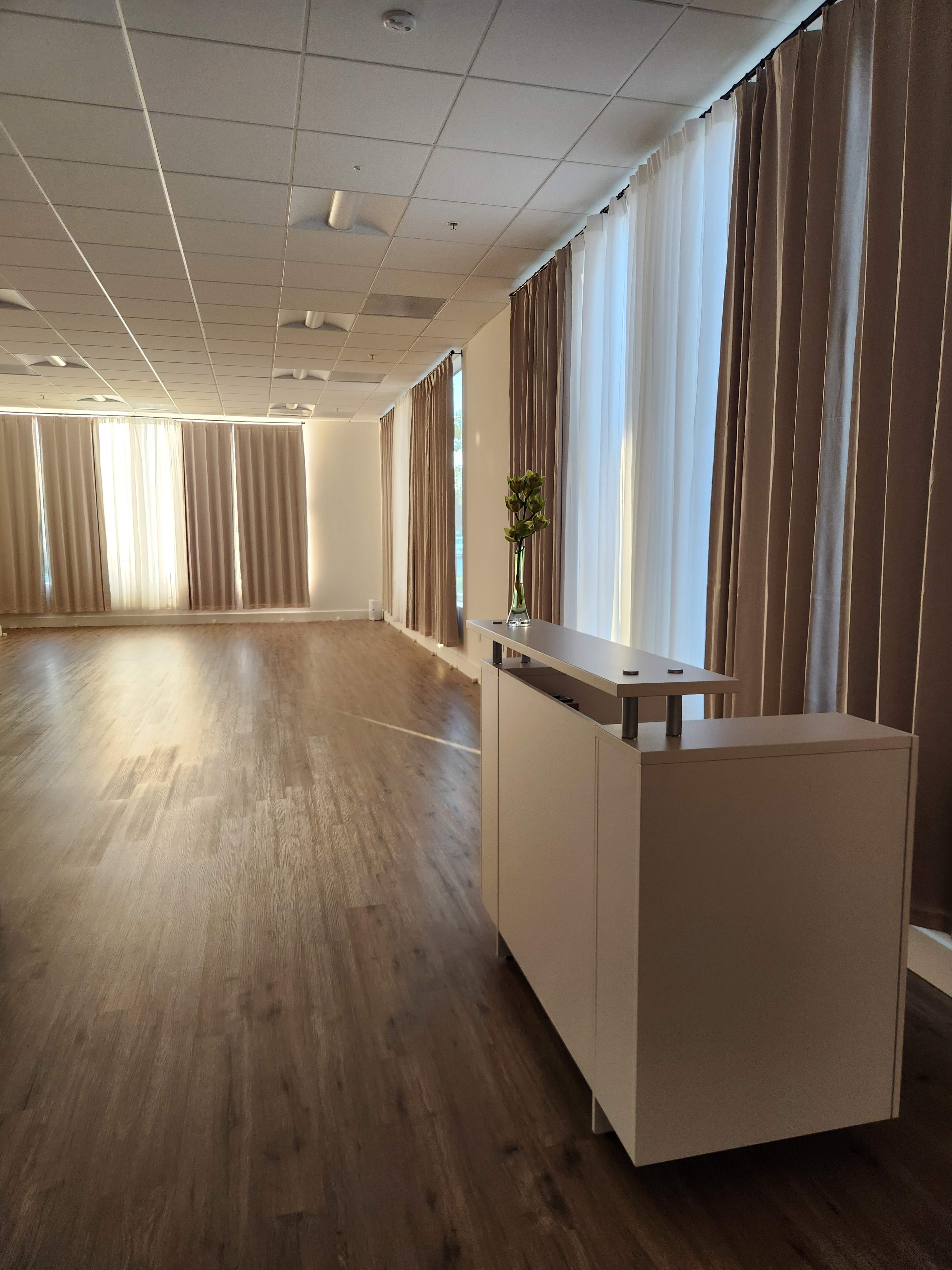 The image shows a spacious room with wooden flooring, large windows covered by beige curtains, and a white console table positioned to one side.