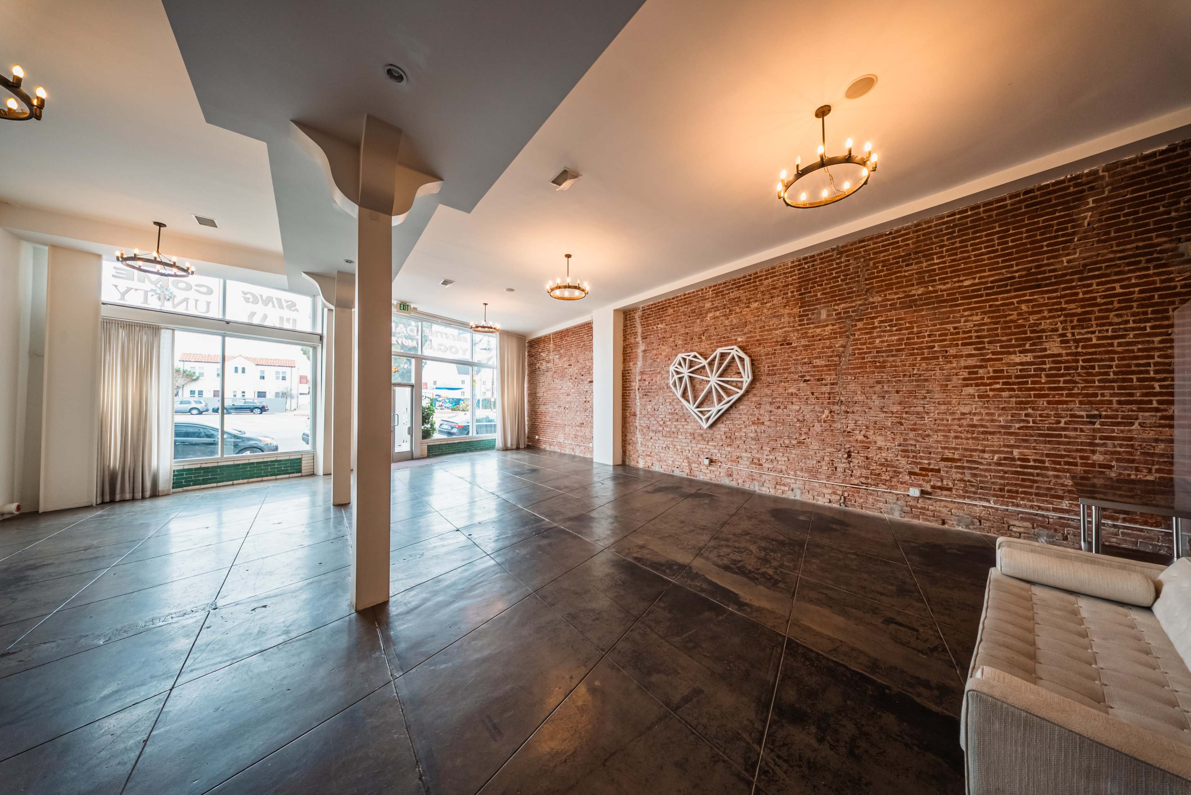The image shows an empty room with exposed brick walls, large windows, and modern chandeliers, featuring a heart-shaped decoration on one wall.
