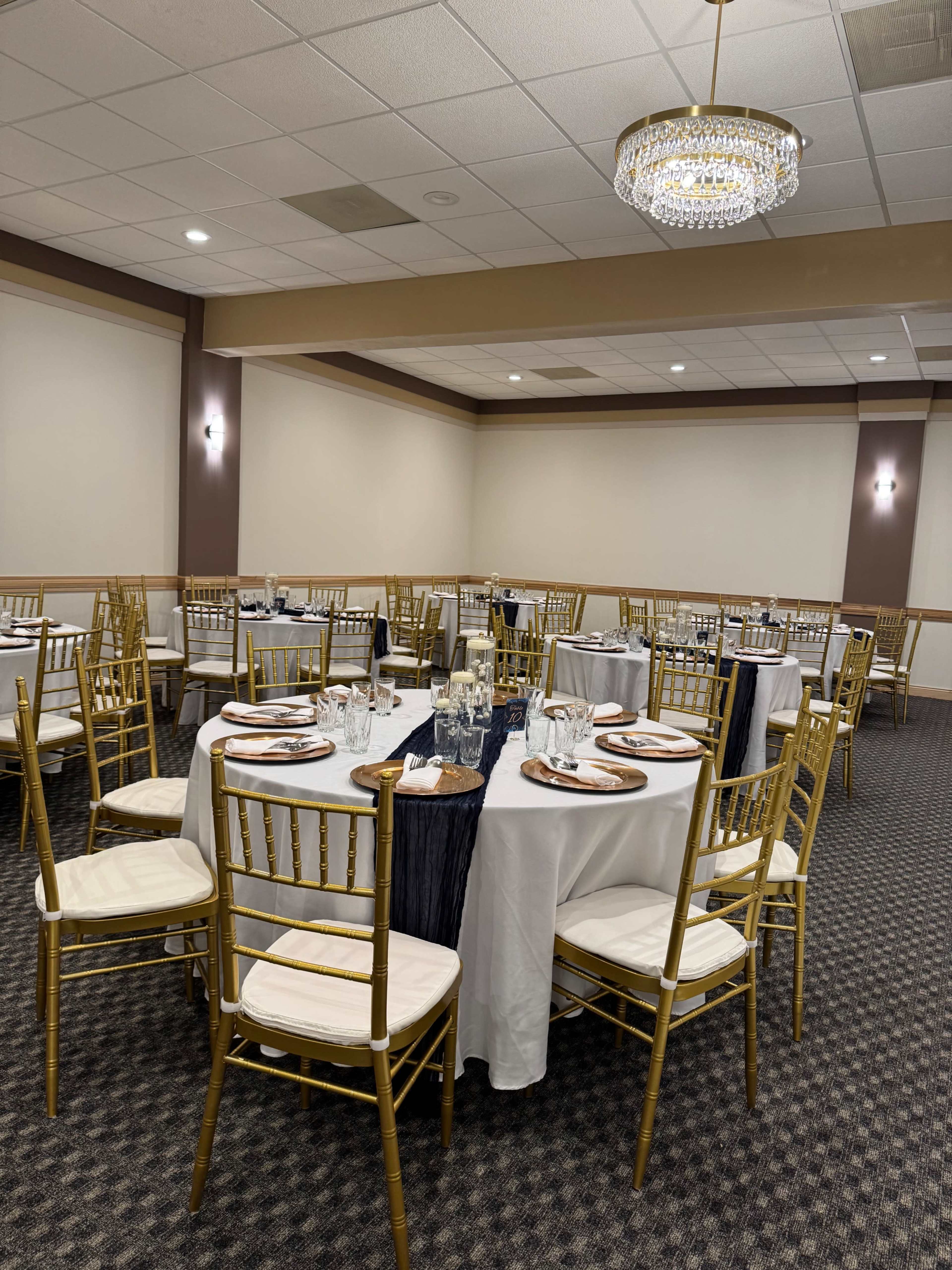 The image shows a dining area with several round tables set with white tablecloths and gold chairs, illuminated by a chandelier overhead.