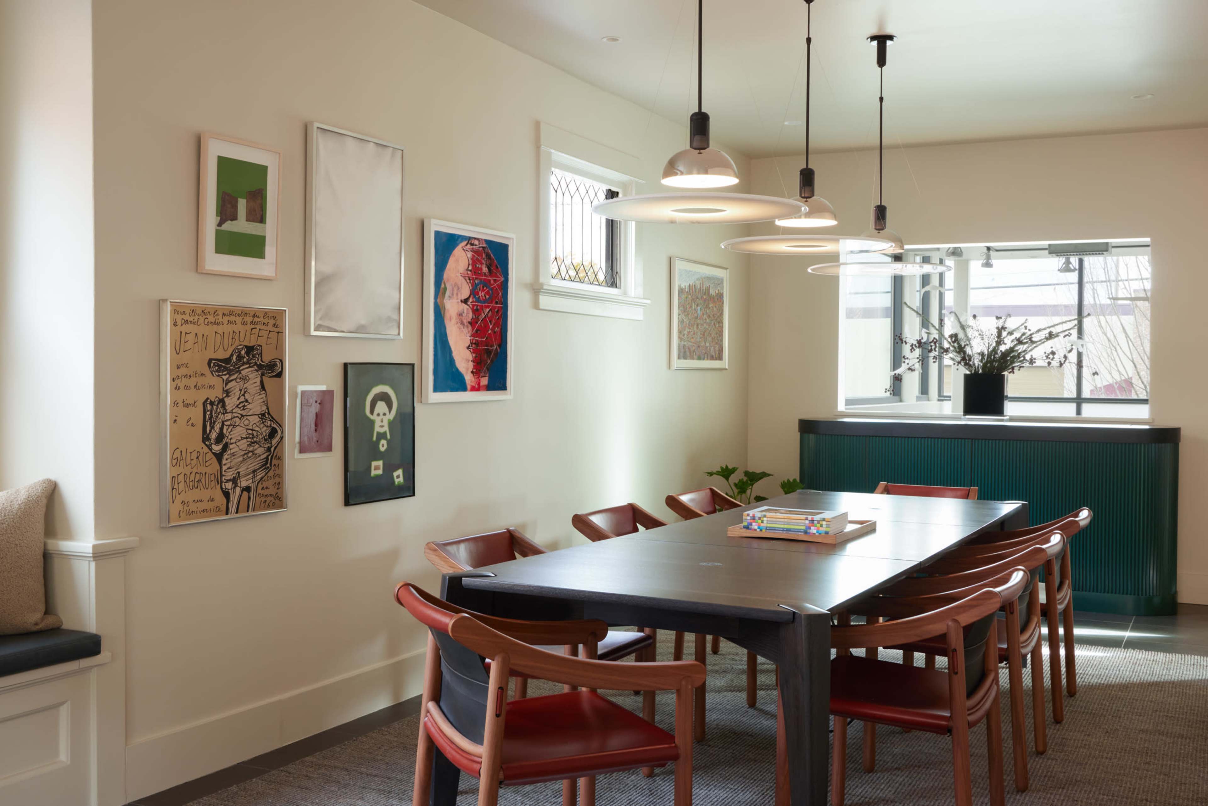 A modern dining room features a long wooden table surrounded by brown chairs, with artwork displayed on the walls and a large window providing natural light.
