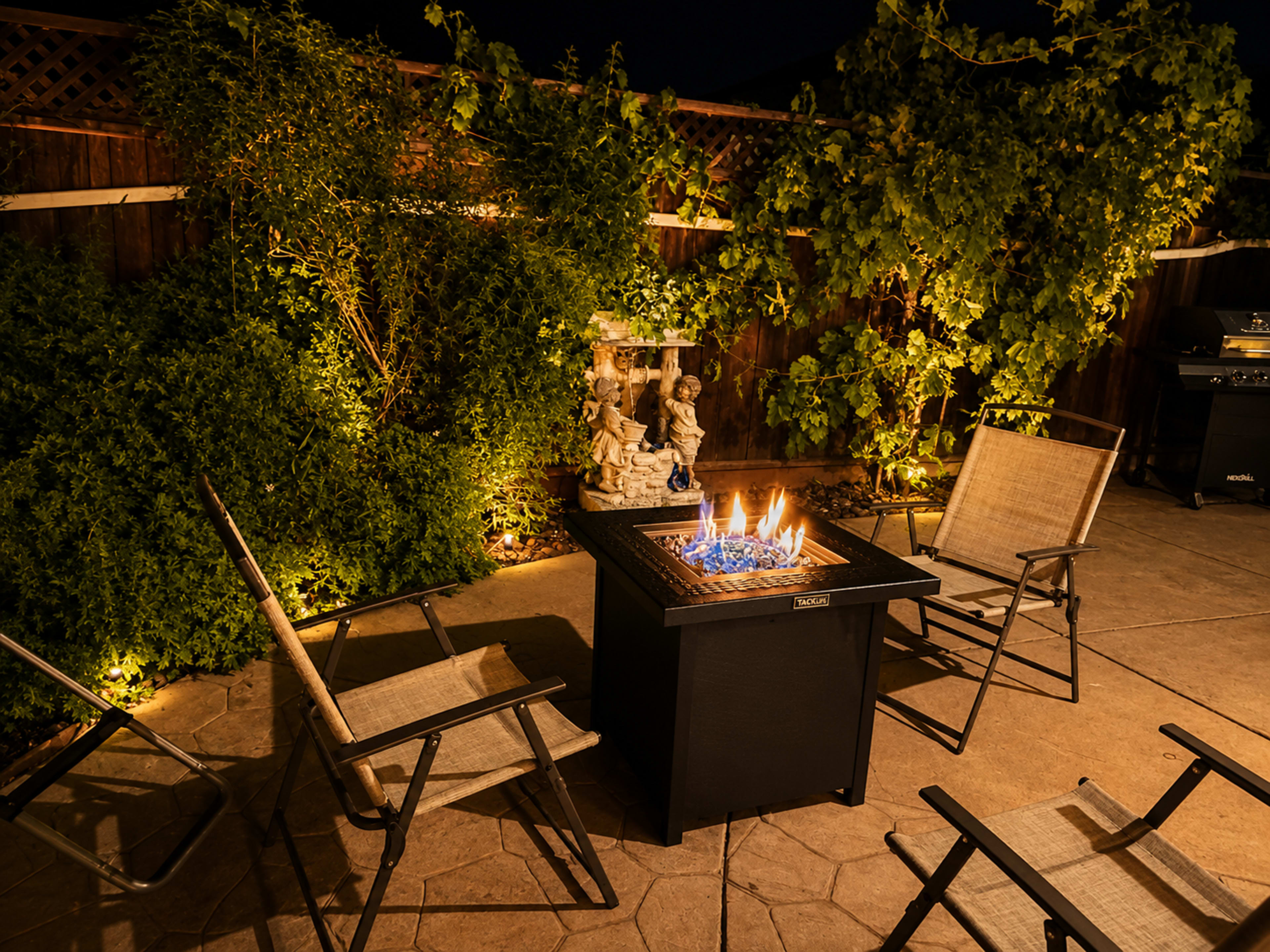 A fire pit surrounded by four chairs is illuminated at night in a garden setting with greenery and soft lighting.