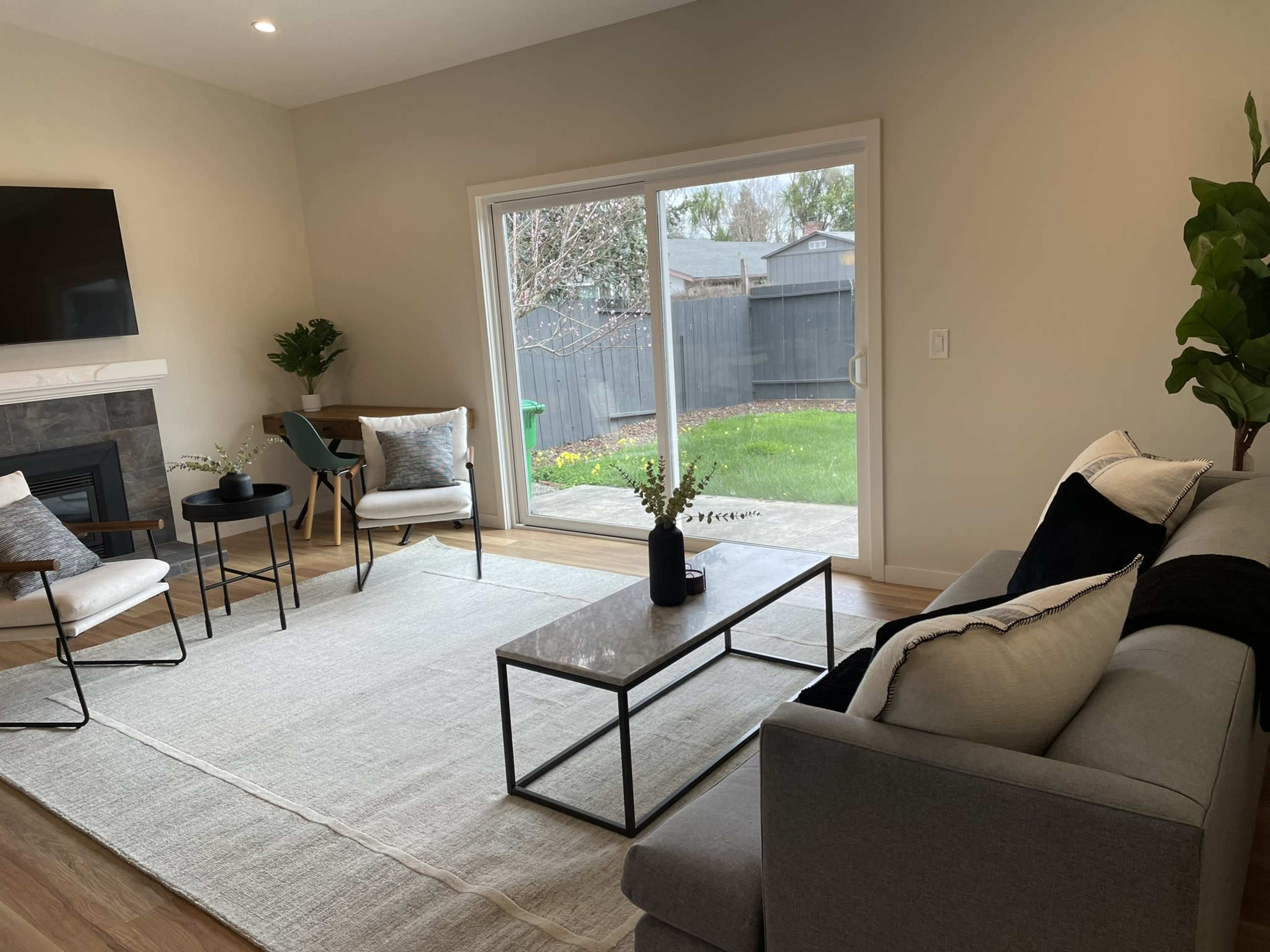 A modern living room with a gray sofa, a coffee table, and two chairs facing a large sliding glass door that opens to a grassy backyard.