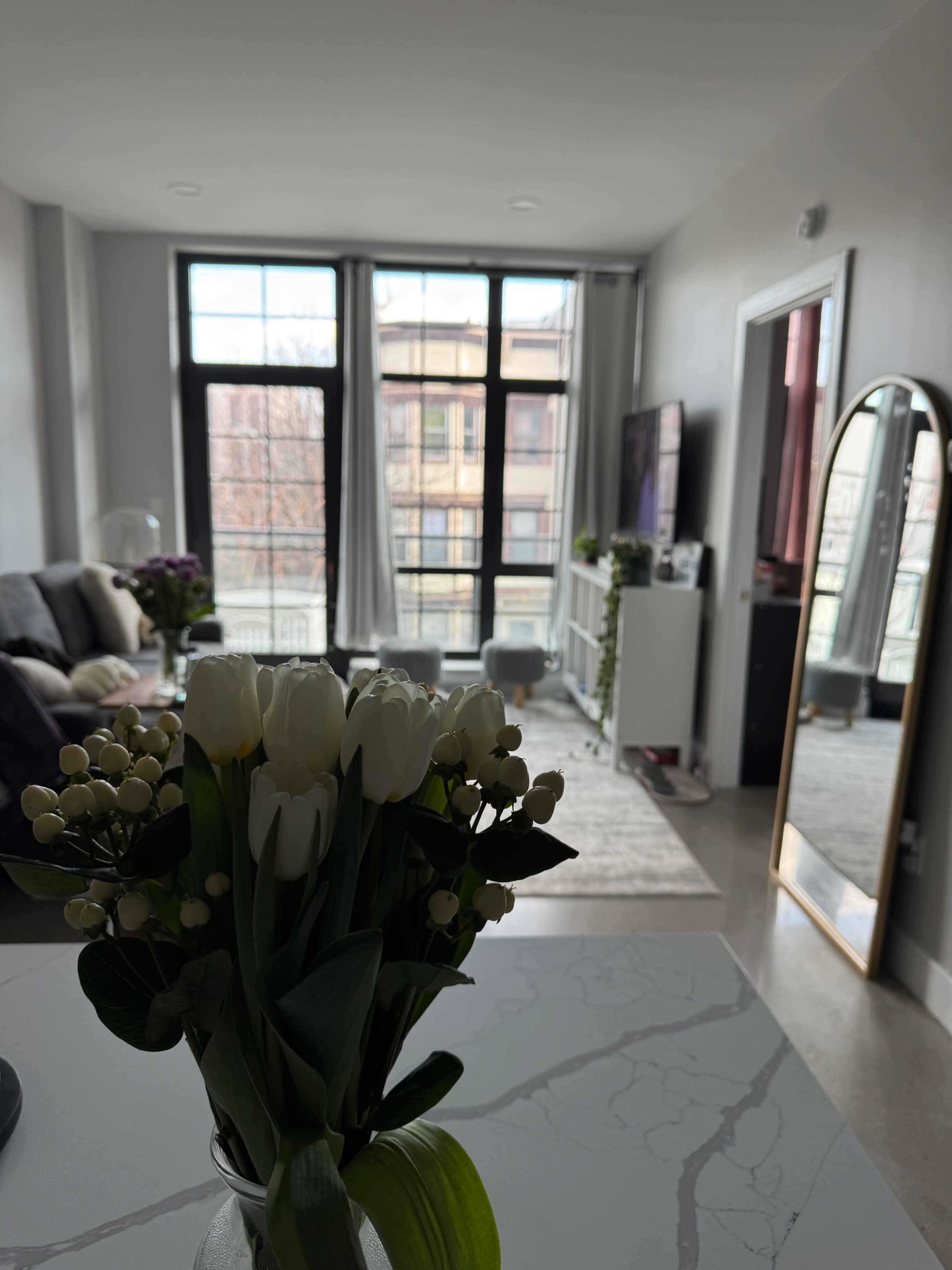 A vase of white tulips is placed in the foreground of a modern living room with large windows and a television.