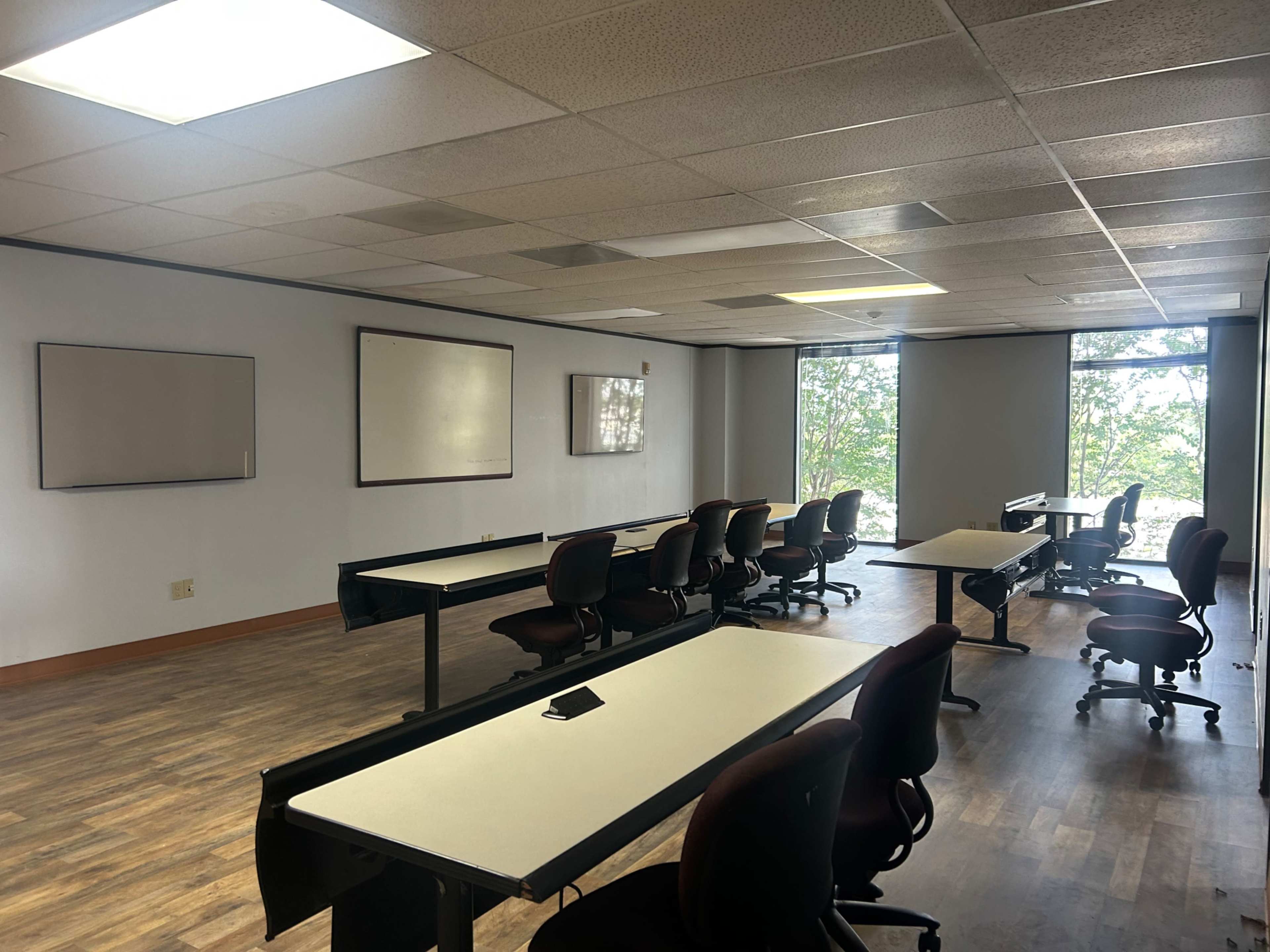 The image shows a spacious, empty classroom with several tables and chairs arranged neatly, large windows allowing natural light, and whiteboards on the walls.