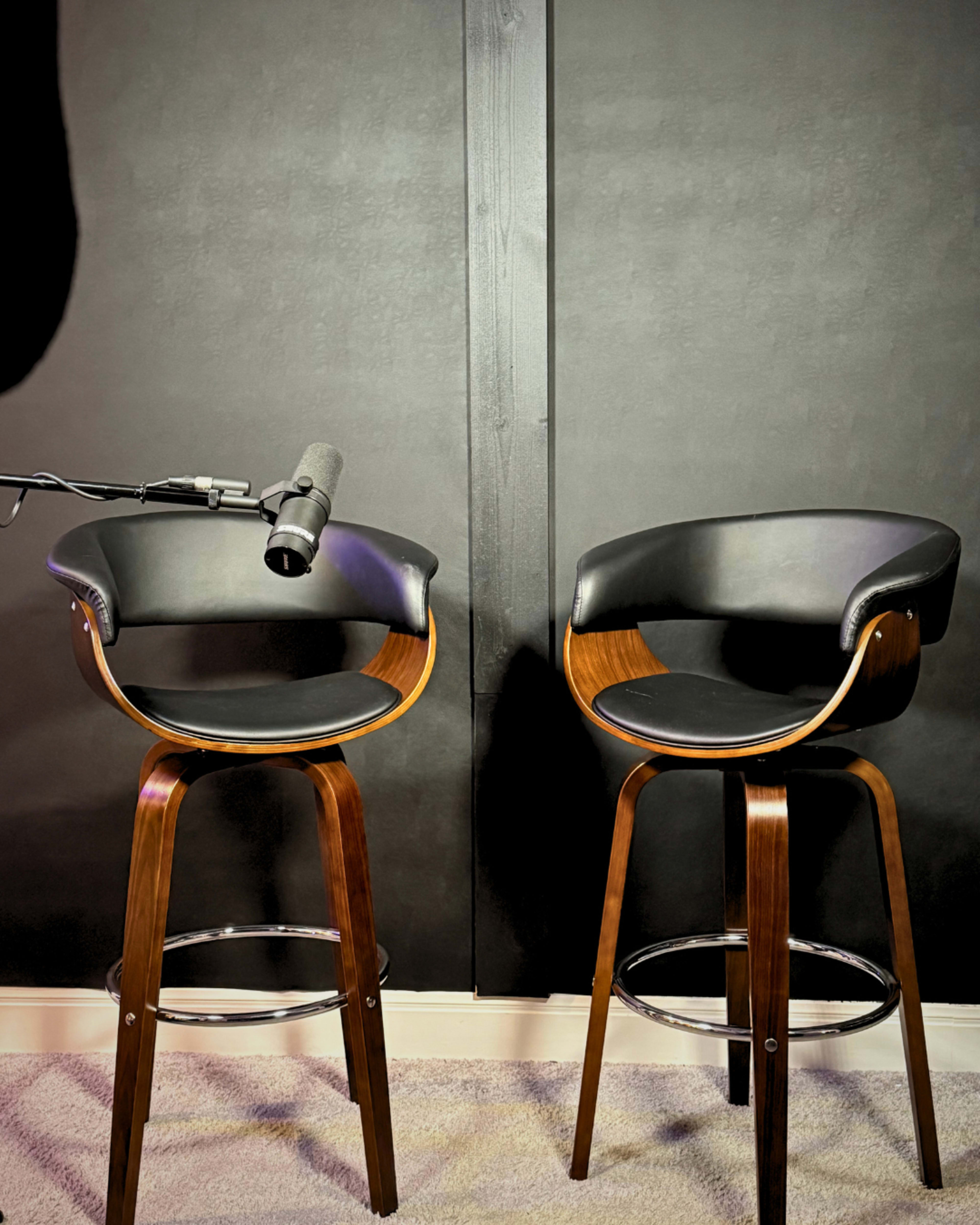 The image shows two black and wooden bar stools positioned against a dark wall, with a microphone mounted on a stand nearby.