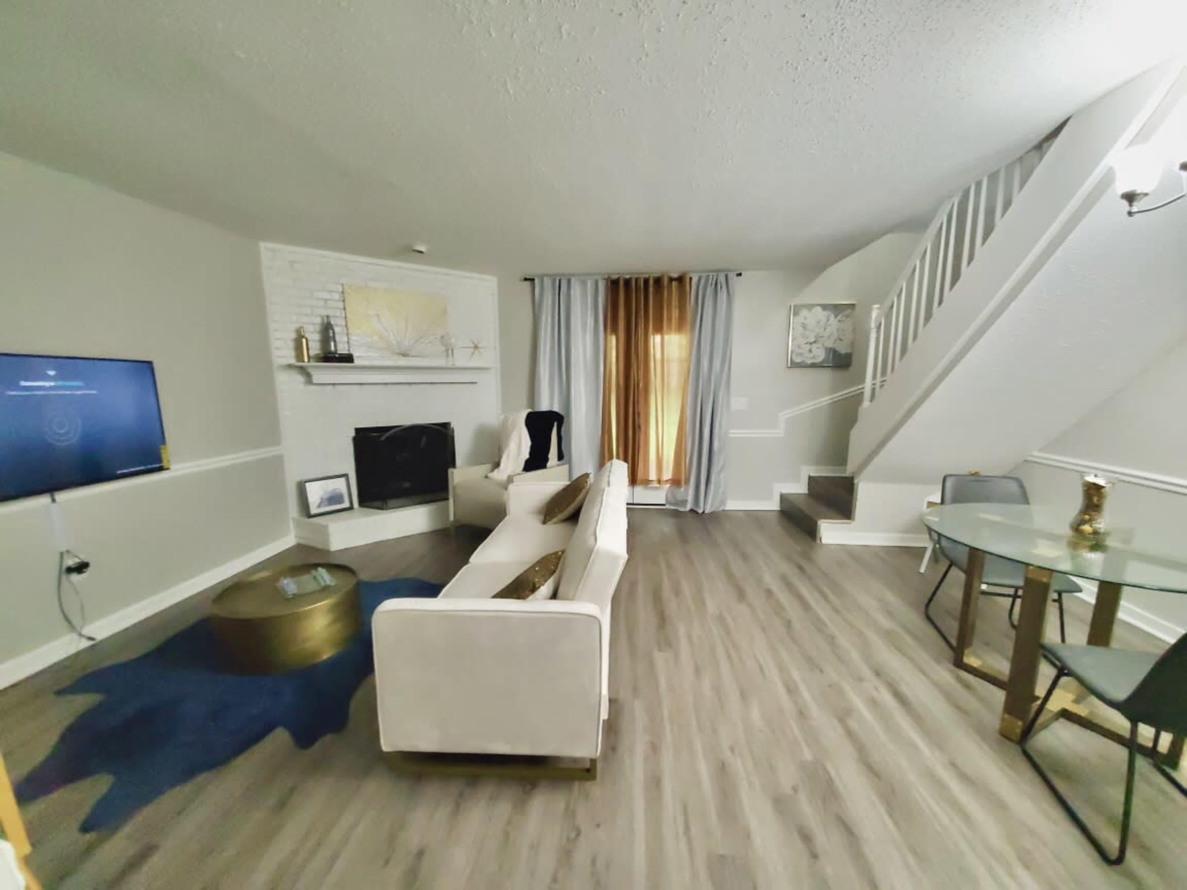 The living room features a white sofa, a television mounted on the wall, a staircase, and a glass dining table accompanied by chairs.