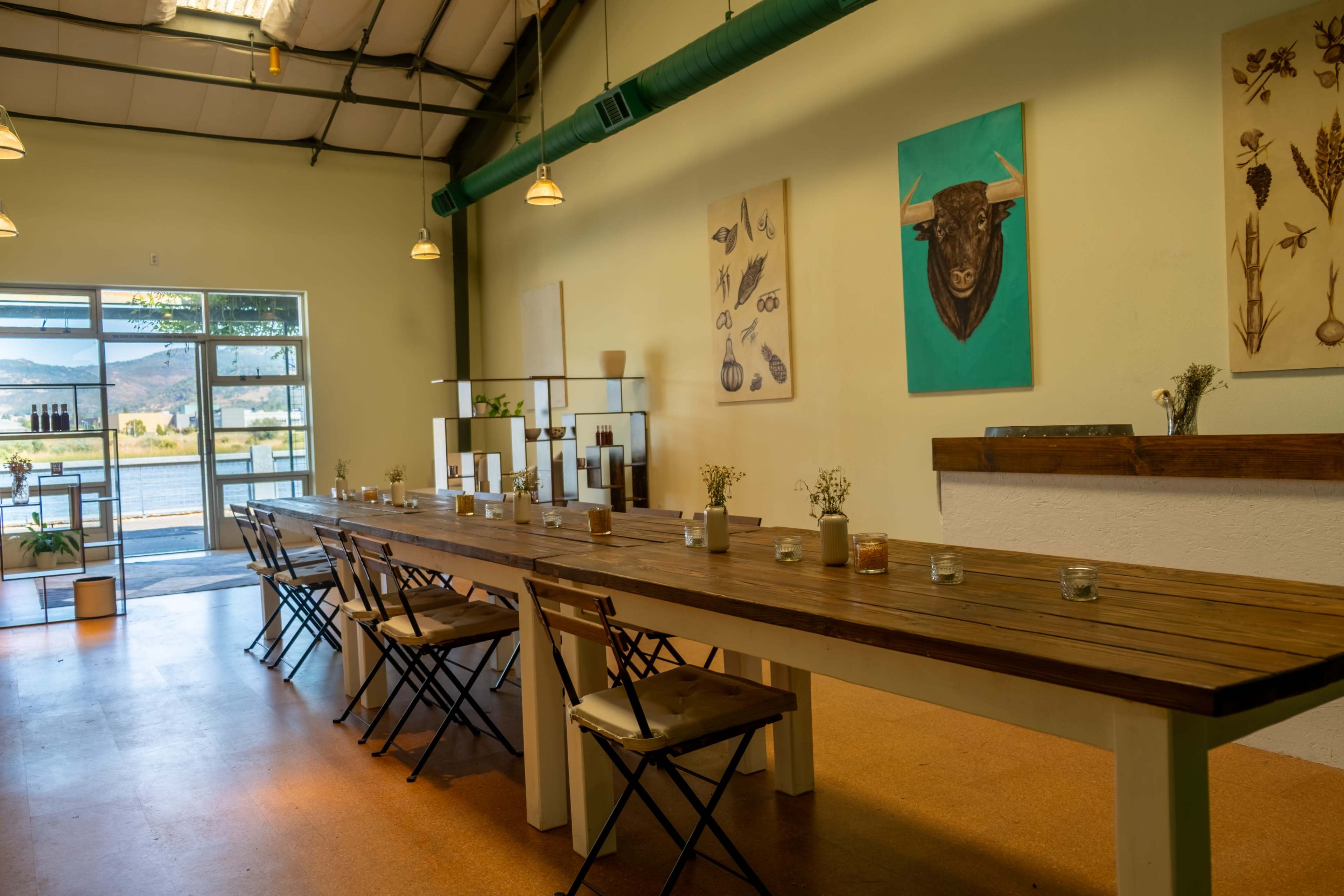 A long wooden table with metal folding chairs is set inside a spacious room featuring large windows and several framed artworks on the walls.