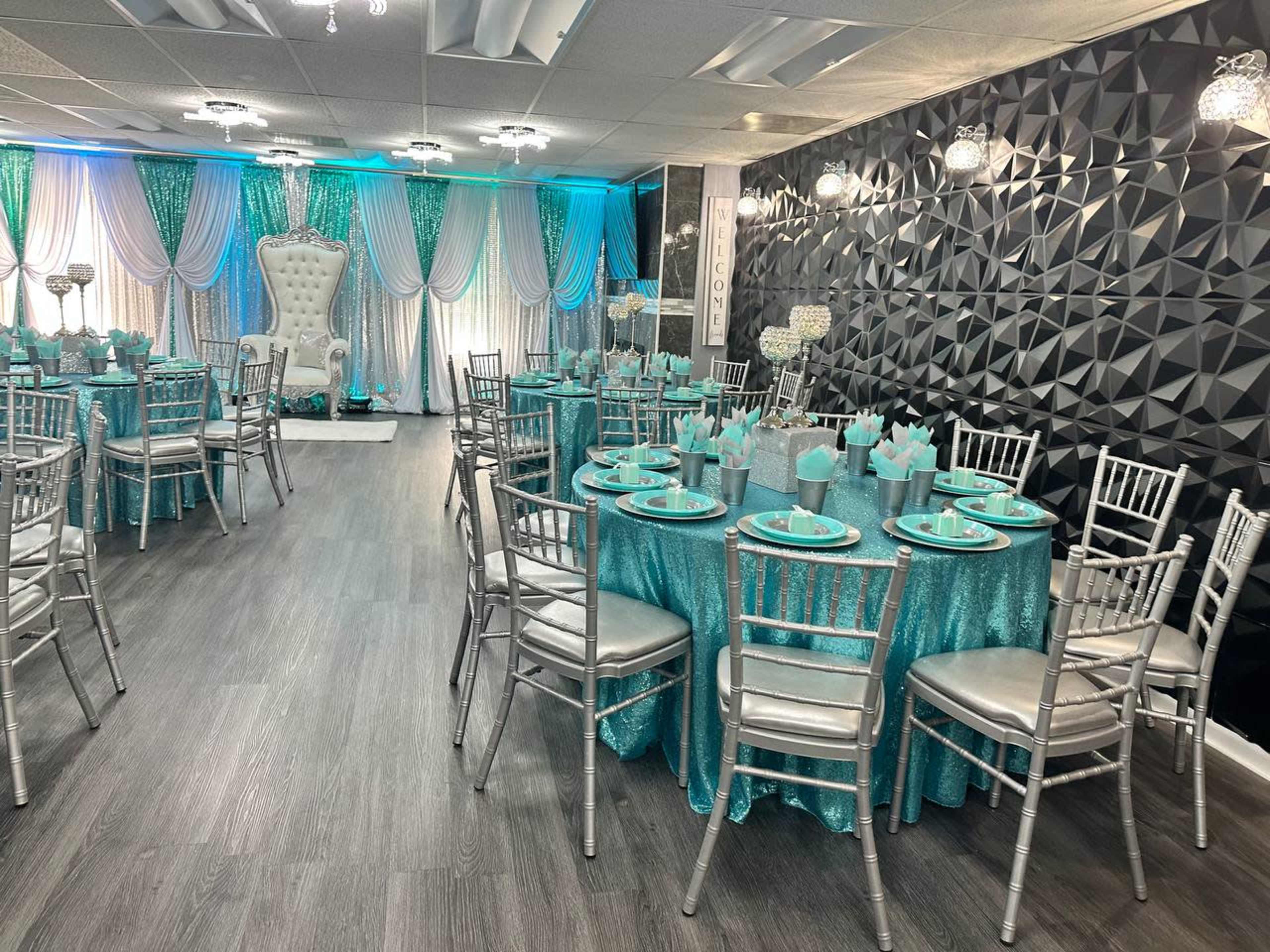 A decorated event space featuring silver chairs and tables draped in turquoise tablecloths, with elegant lighting and a large white throne at one end.