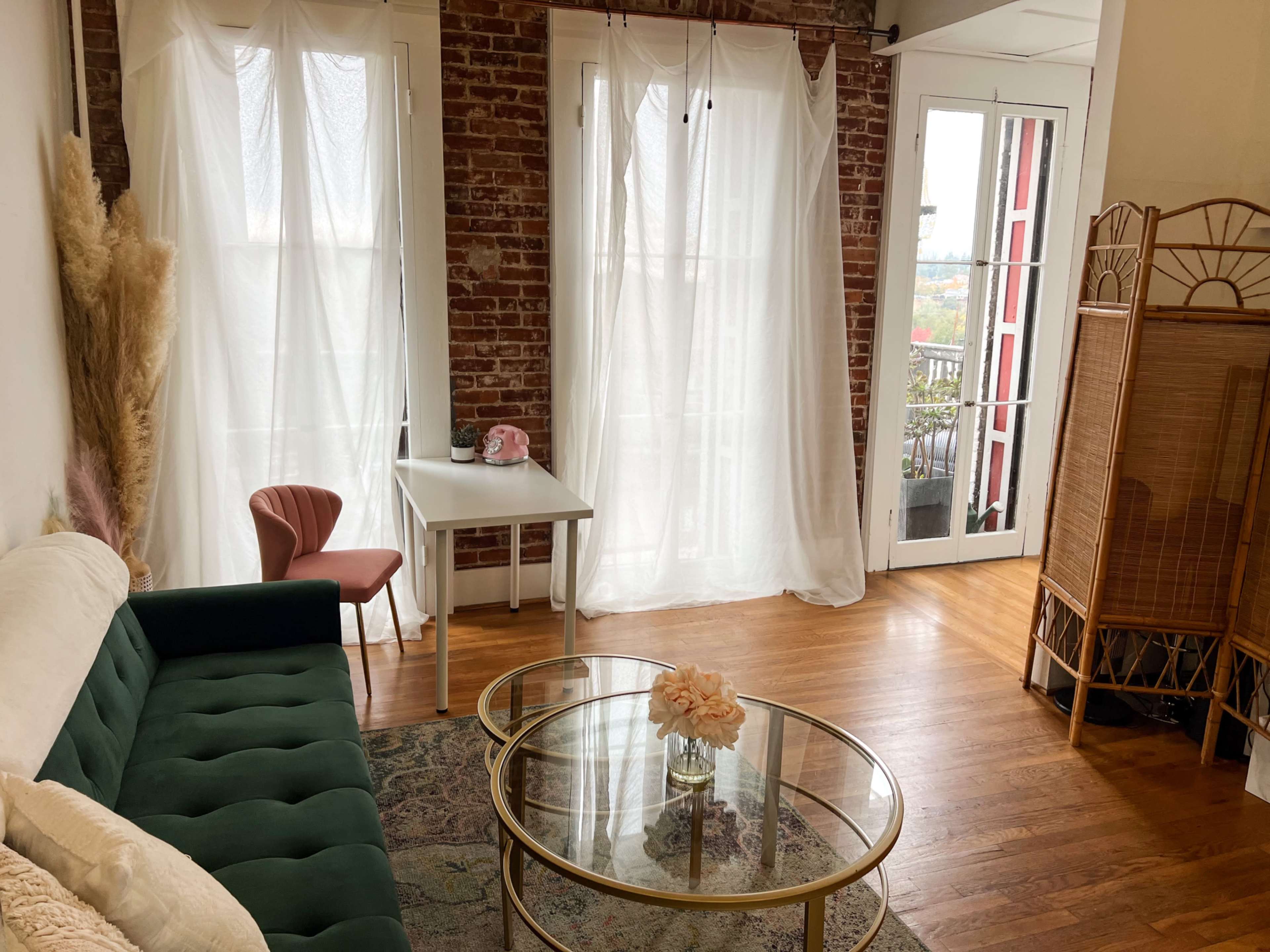 The image shows a bright living space with a green sofa, a glass coffee table, a small white table, and large windows dressed with sheer curtains.