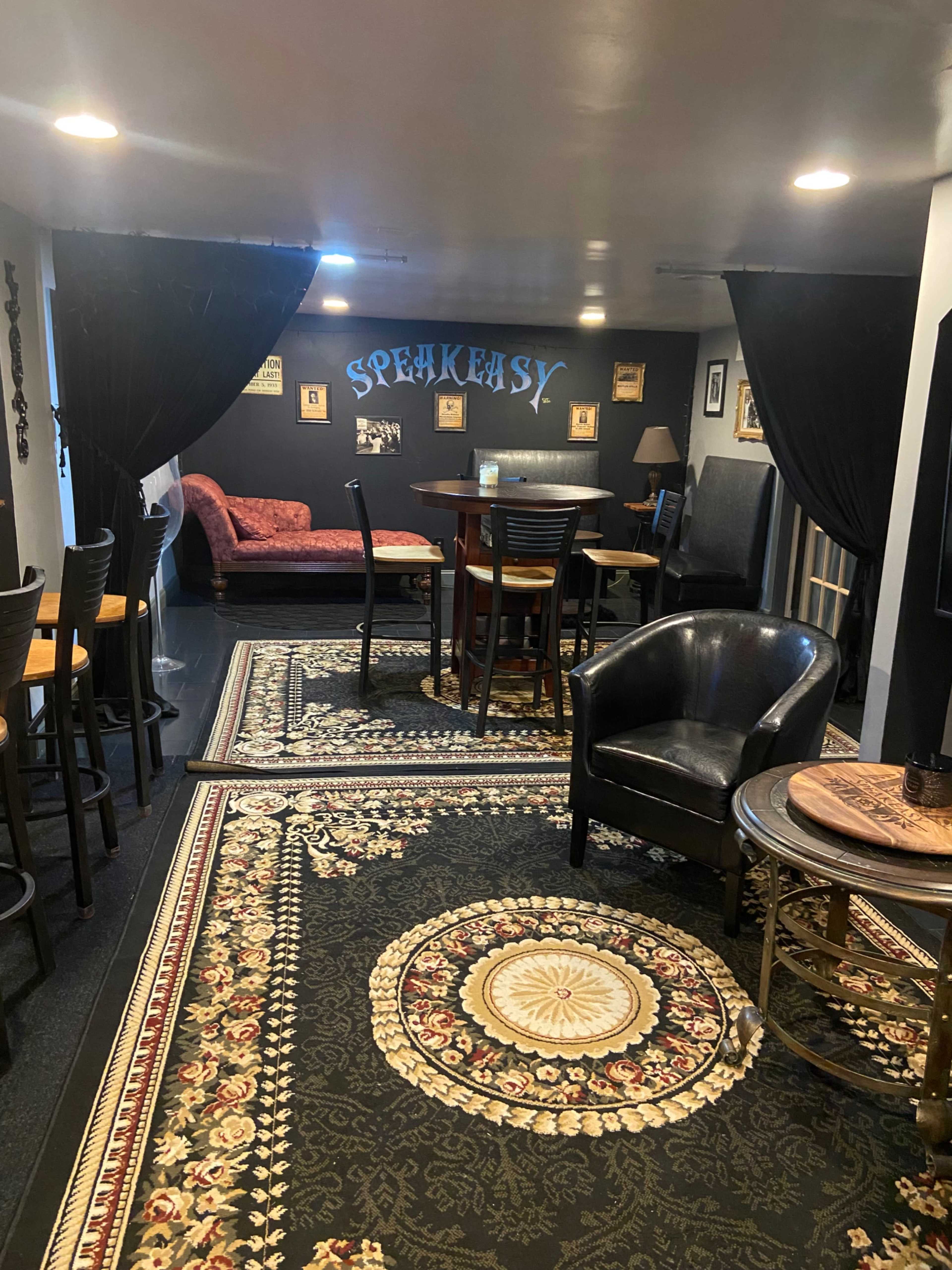 A basement lounge area labeled "SPEAKEASY," featuring a patterned rug, a seating arrangement with chairs and a small table, and decorative wall art.