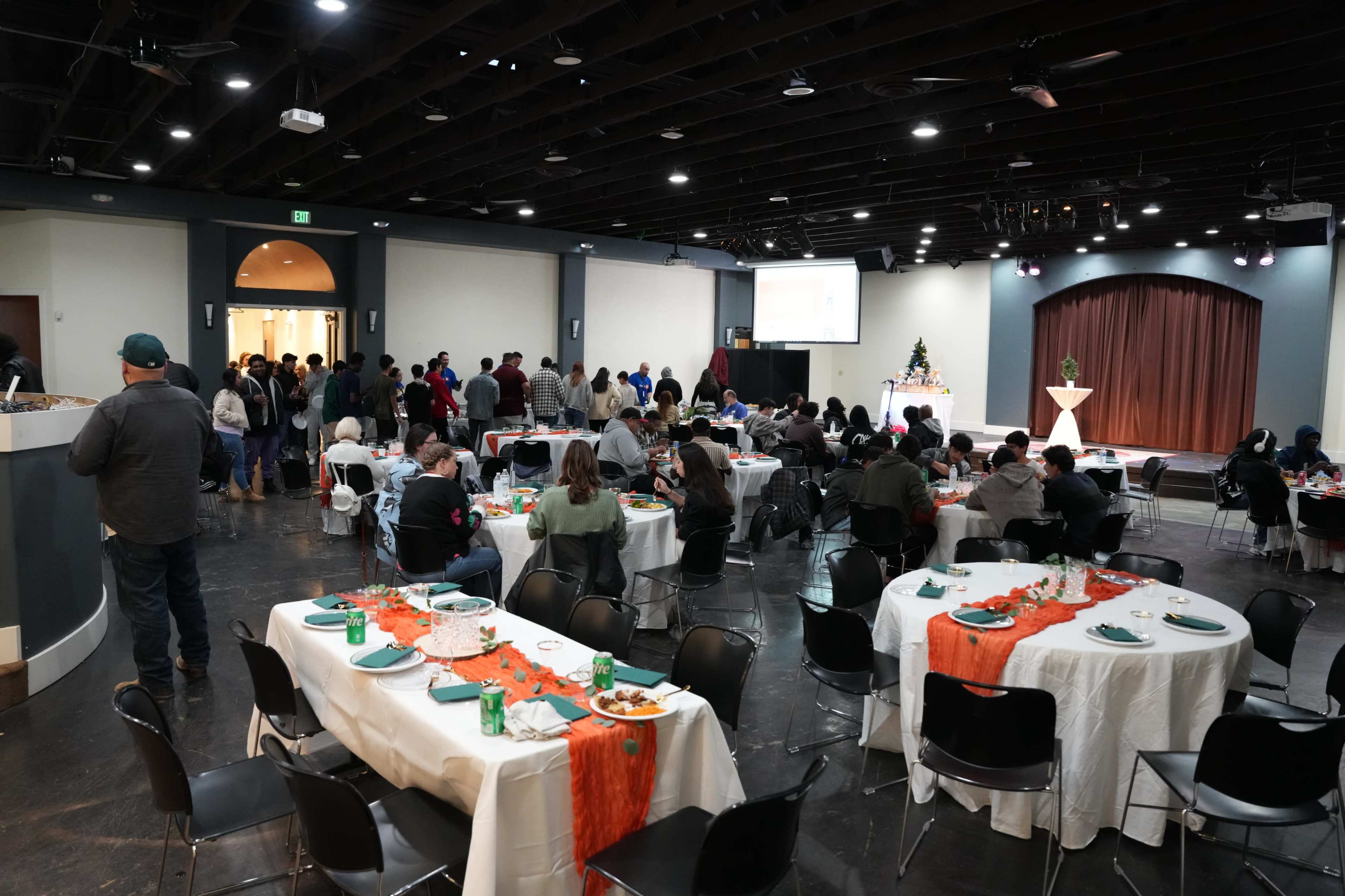 A large group of people gathers in a banquet hall with tables set for a meal and a stage at the front.