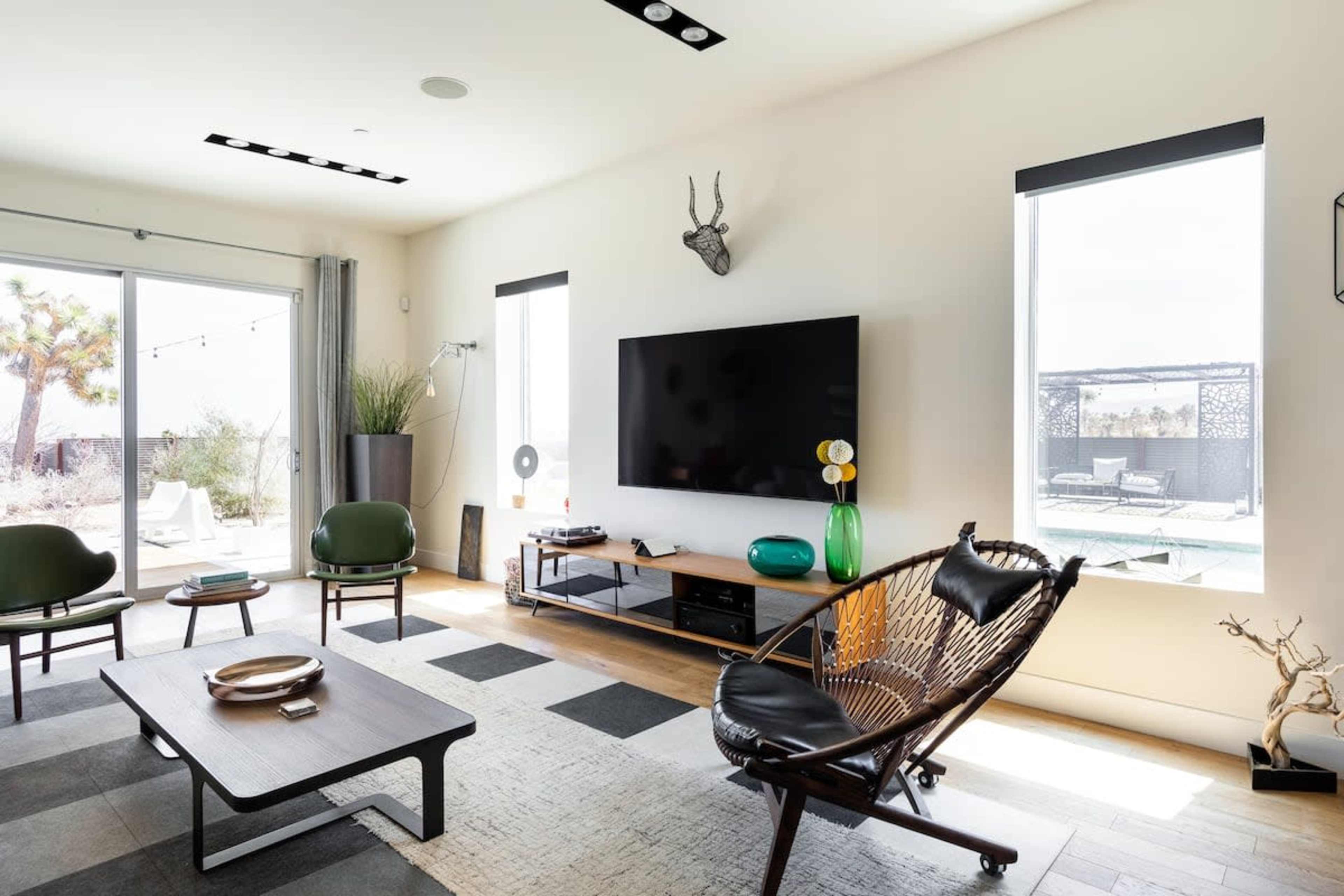 The living room features a television mounted on a wall above a wooden media console, with seating consisting of a round black chair and two green chairs, set against large windows that open to a patio.