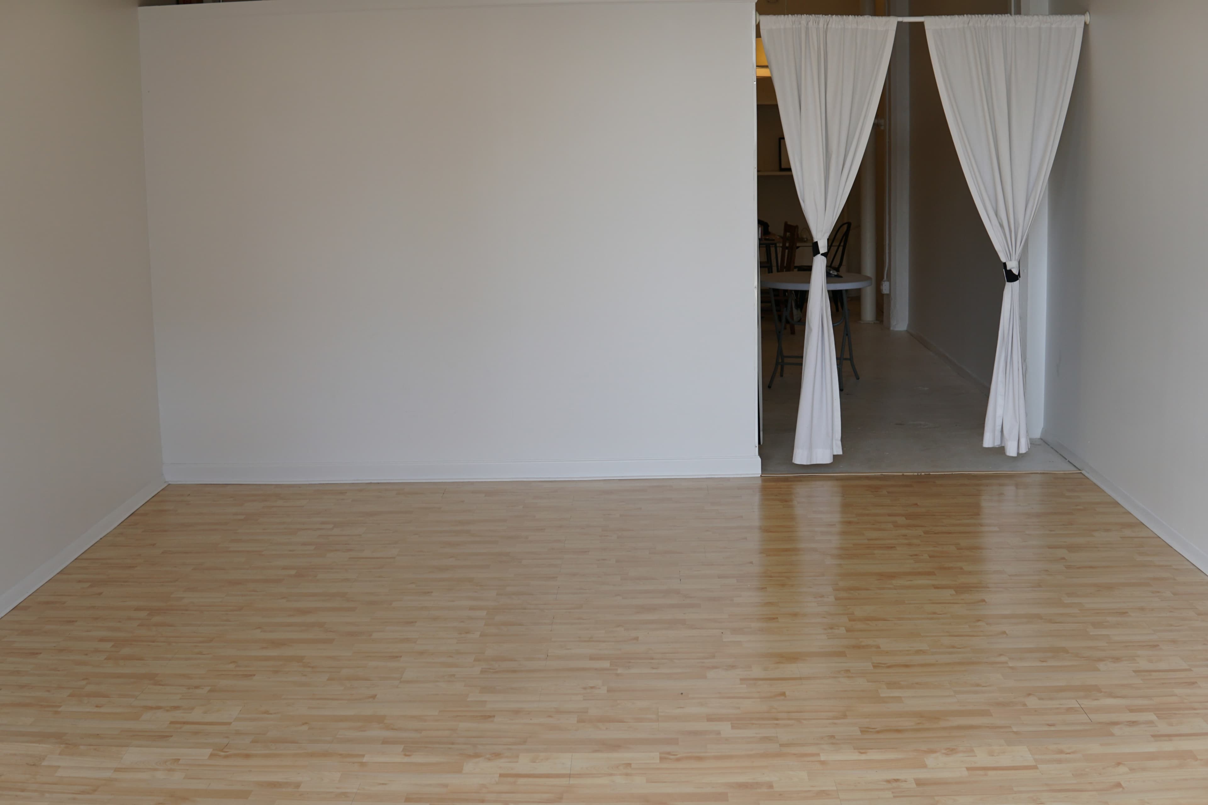 A spacious room with a light wooden floor and a doorway draped with white curtains leading to an adjacent area.