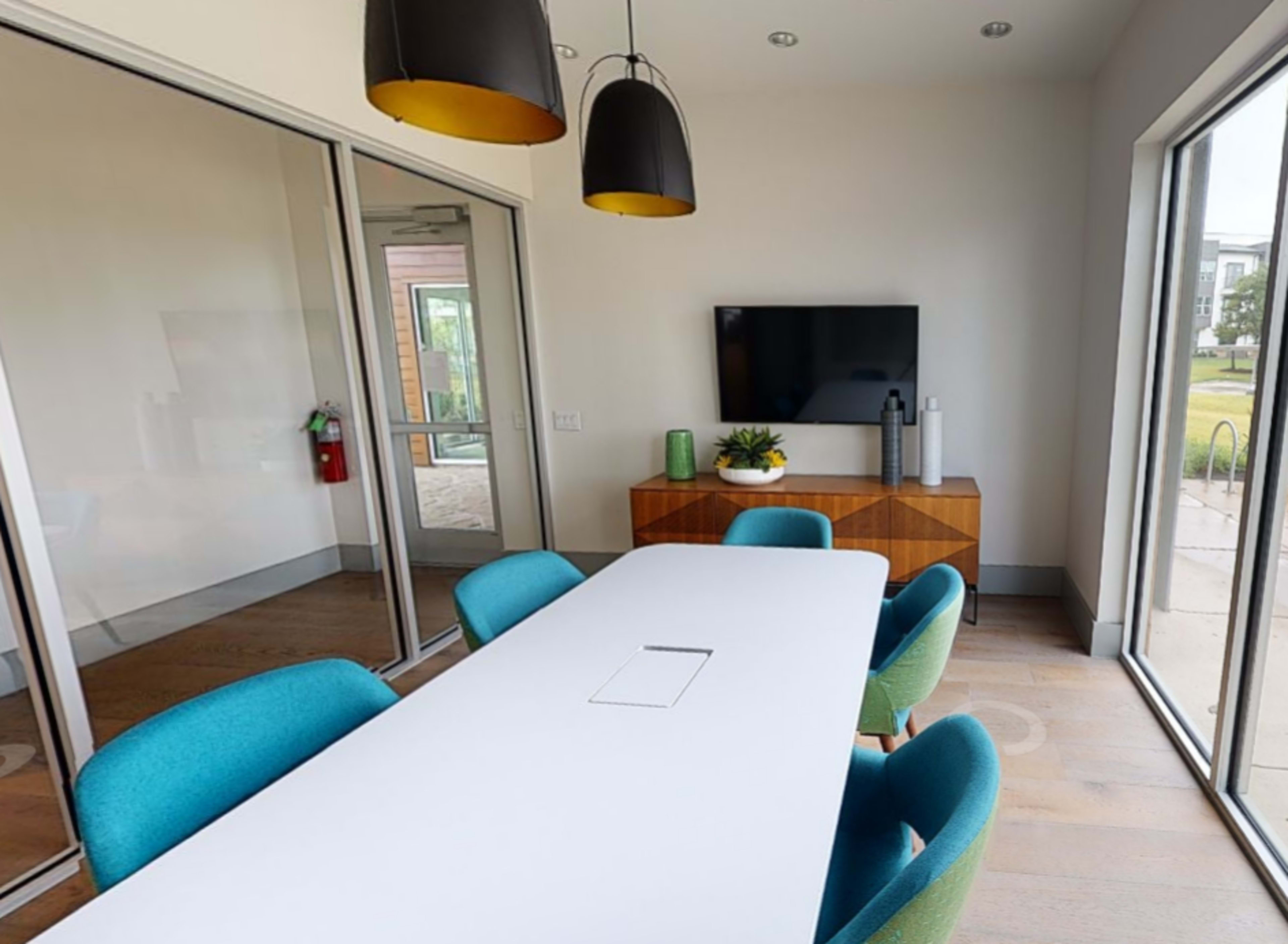 The image shows a modern conference room featuring a long white table, colorful chairs, a television mounted on the wall, and large windows allowing natural light.