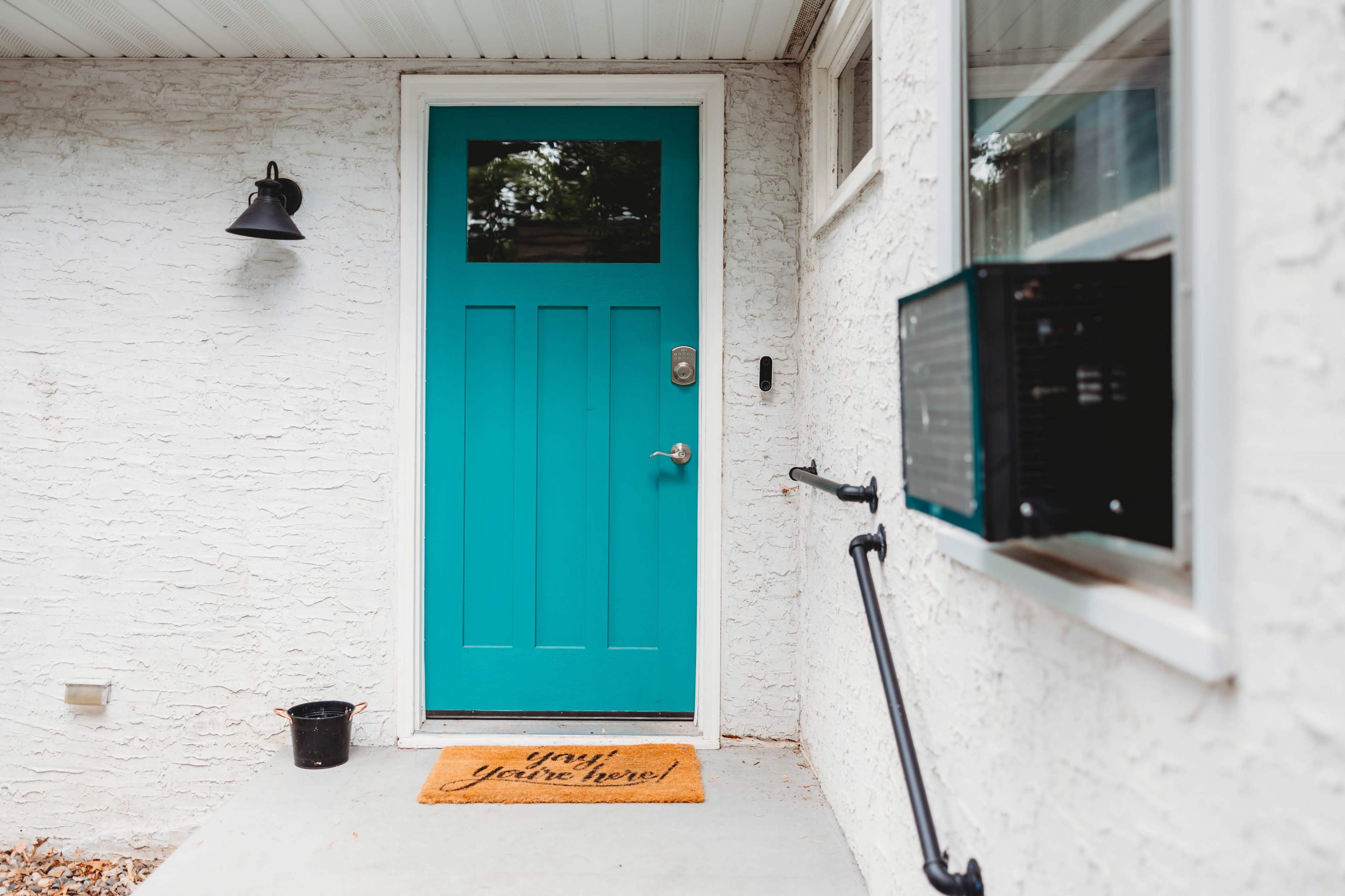 A teal front door with a welcome mat is set against a white textured wall, next to a window and an air conditioning unit.