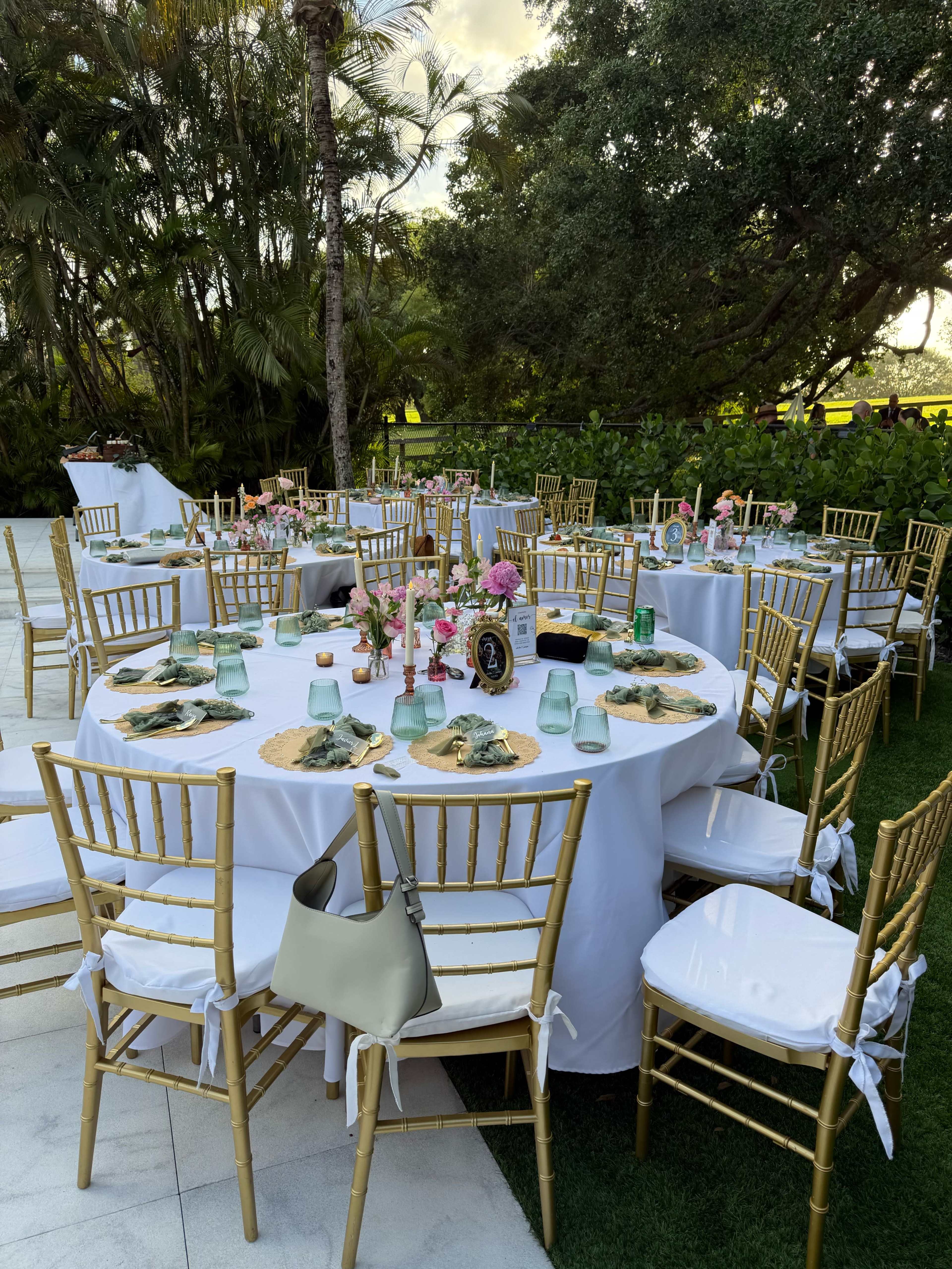 The image shows elegantly set round tables with gold chairs and floral centerpieces in a garden setting, surrounded by greenery.