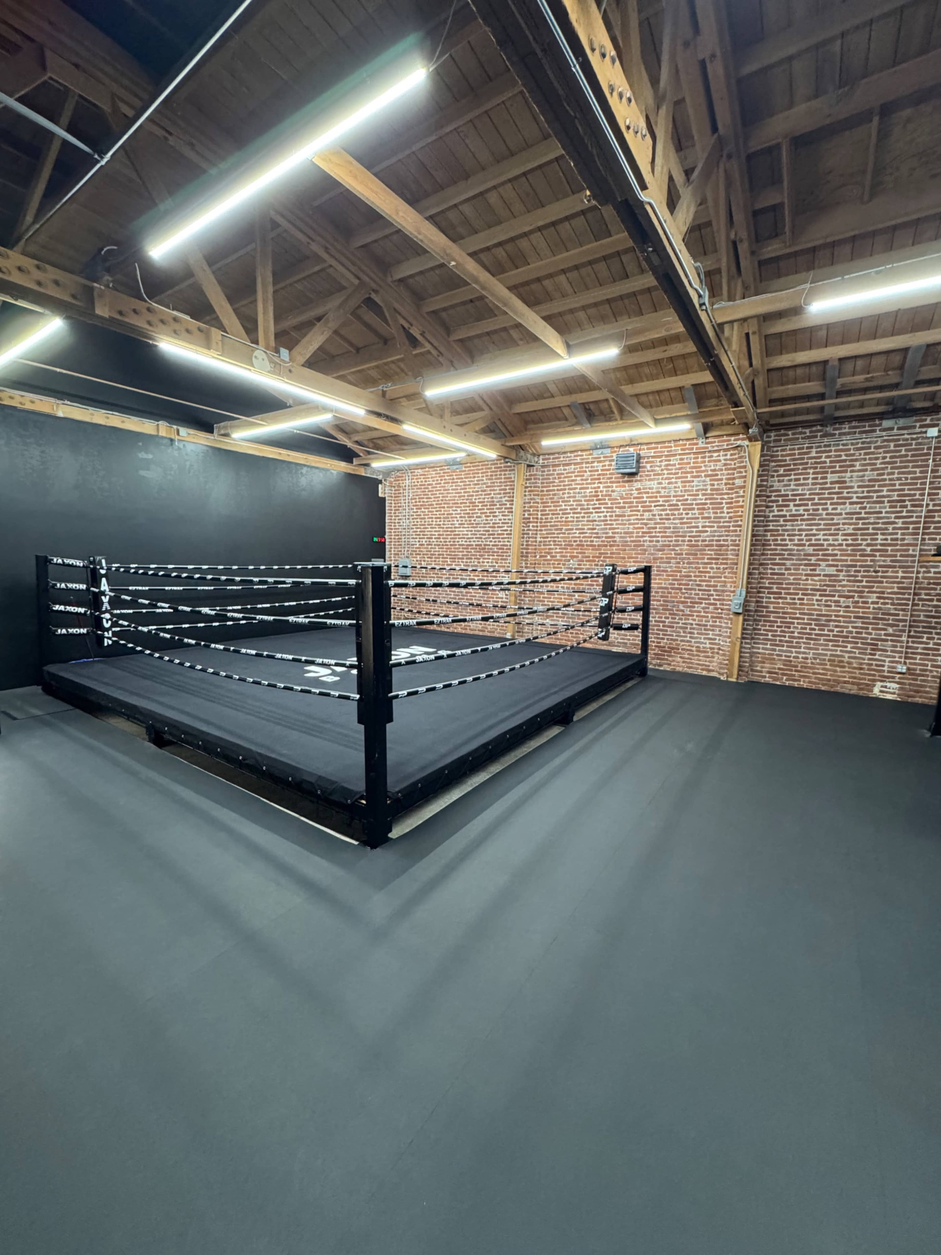 The image shows an empty wrestling ring set up in an industrial-style gym with exposed brick walls and wooden beams overhead.