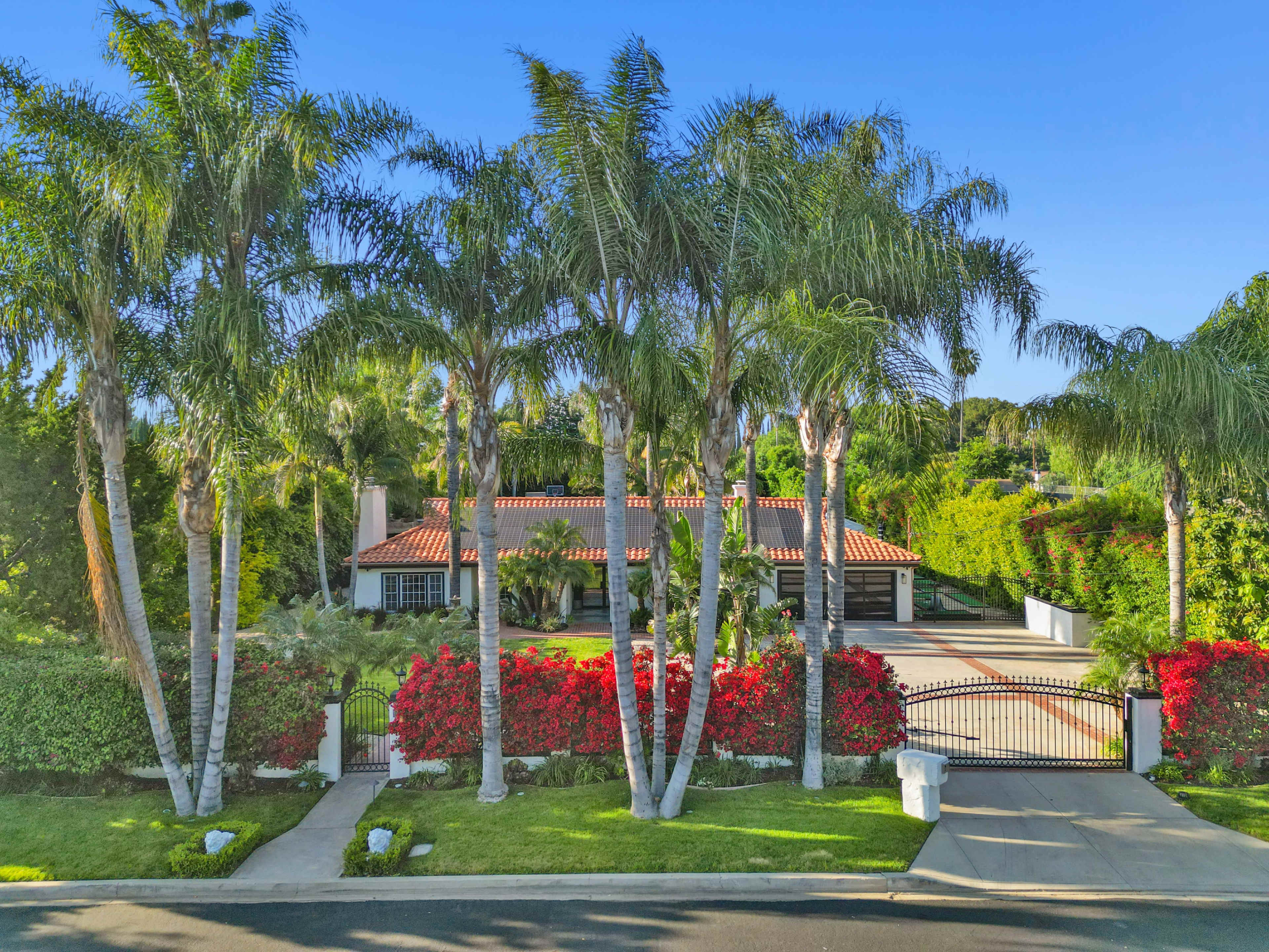A single-story house with a red-tiled roof is surrounded by palm trees and vibrant bougainvillea, set behind a gated entrance.
