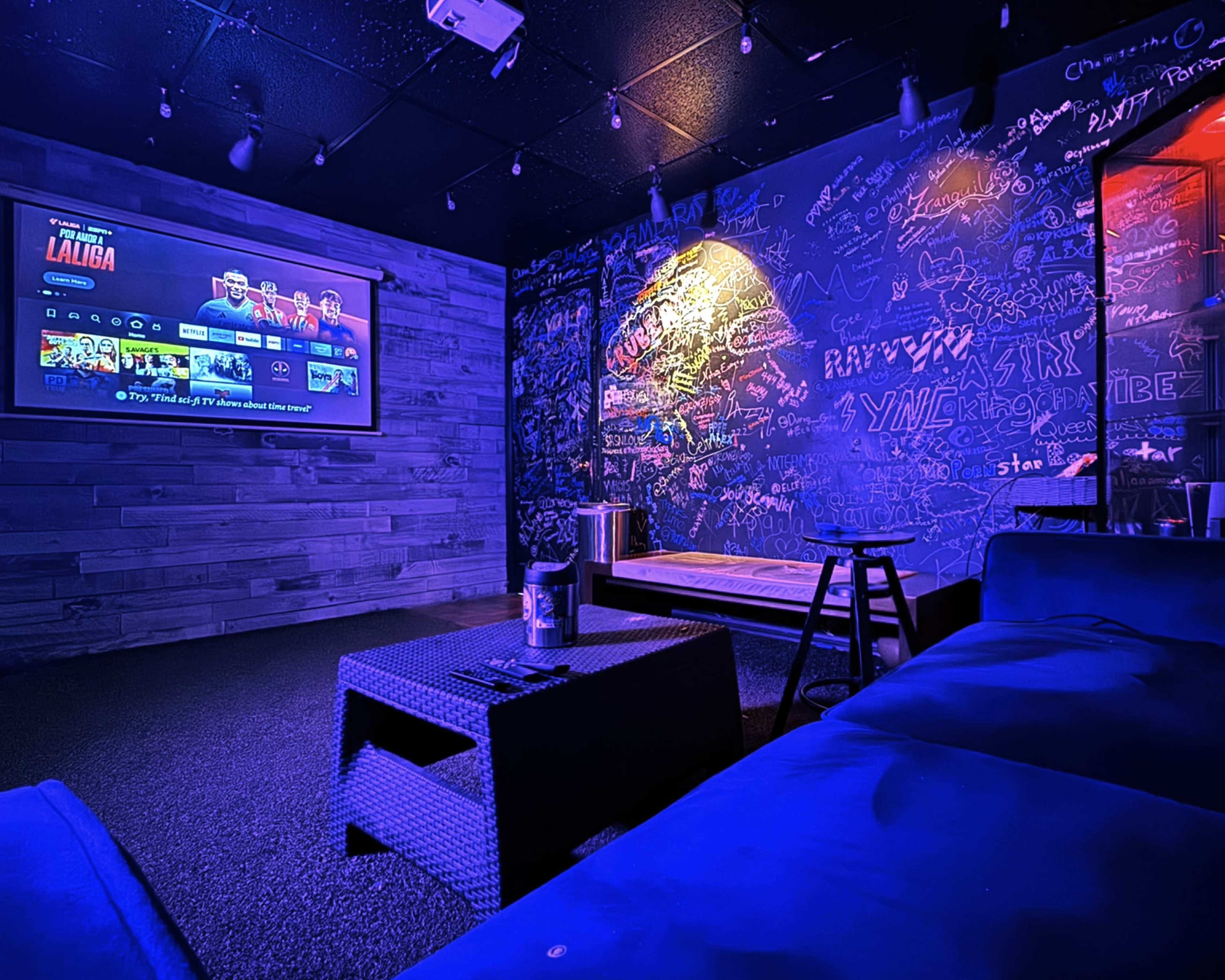 The image features a cozy room with a large screen displaying entertainment options, a couch, a table, and walls covered in colorful graffiti.