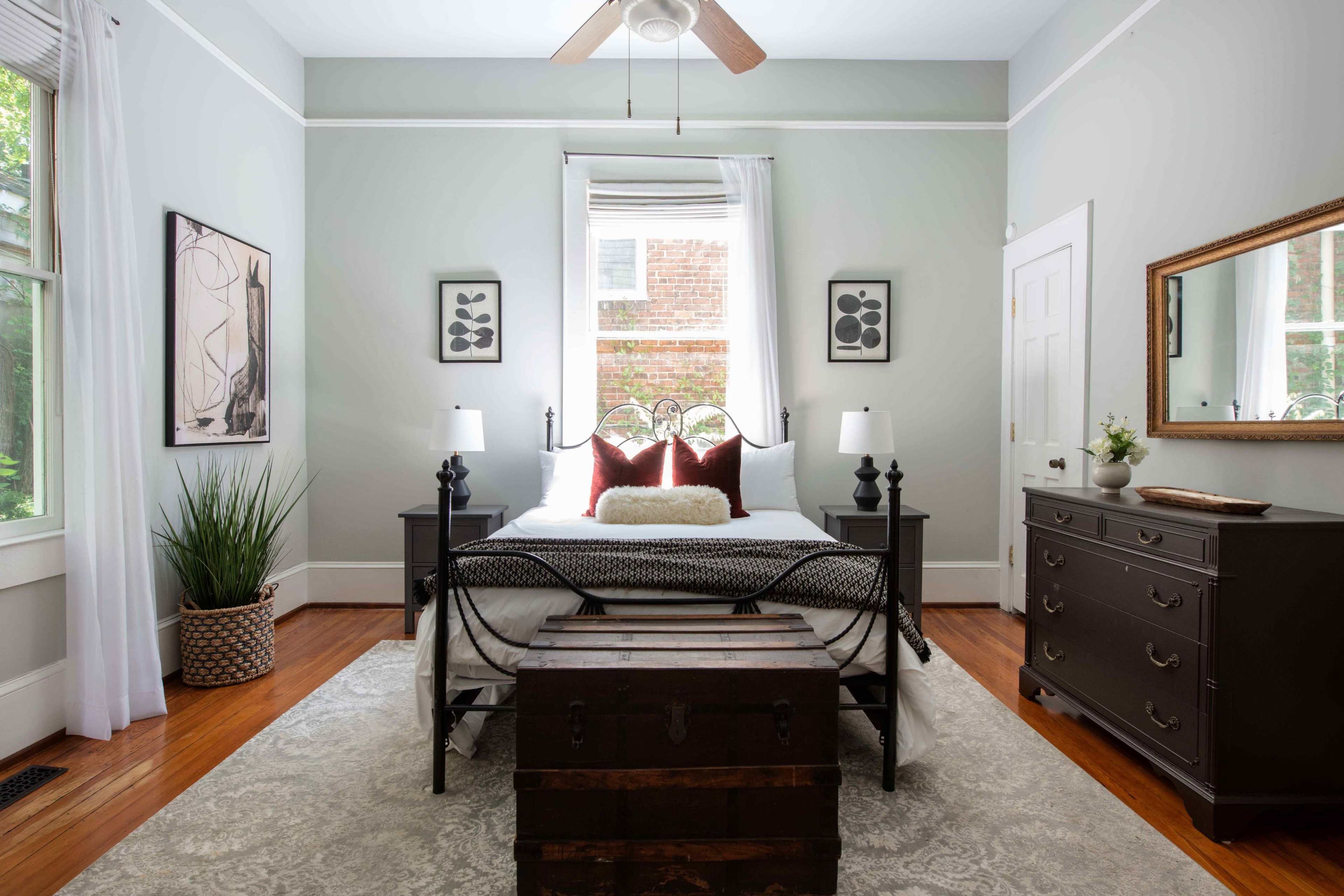 The image shows a well-decorated bedroom featuring a black metal bed frame, two bedside lamps, a wooden dresser, and a trunk at the foot of the bed, all set against light gray walls.