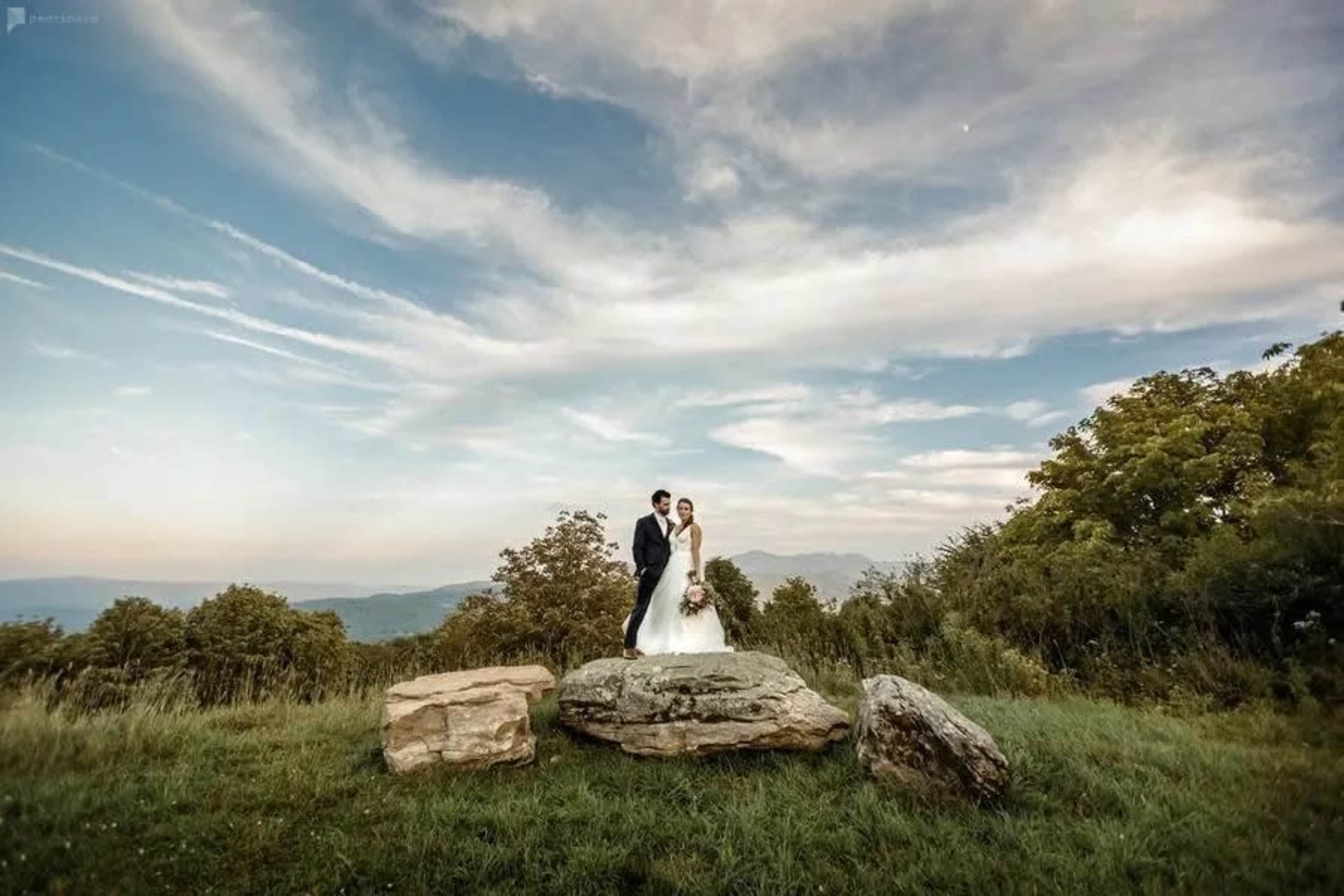 A couple stands on a rocky outcrop surrounded by greenery under a cloudy sky.