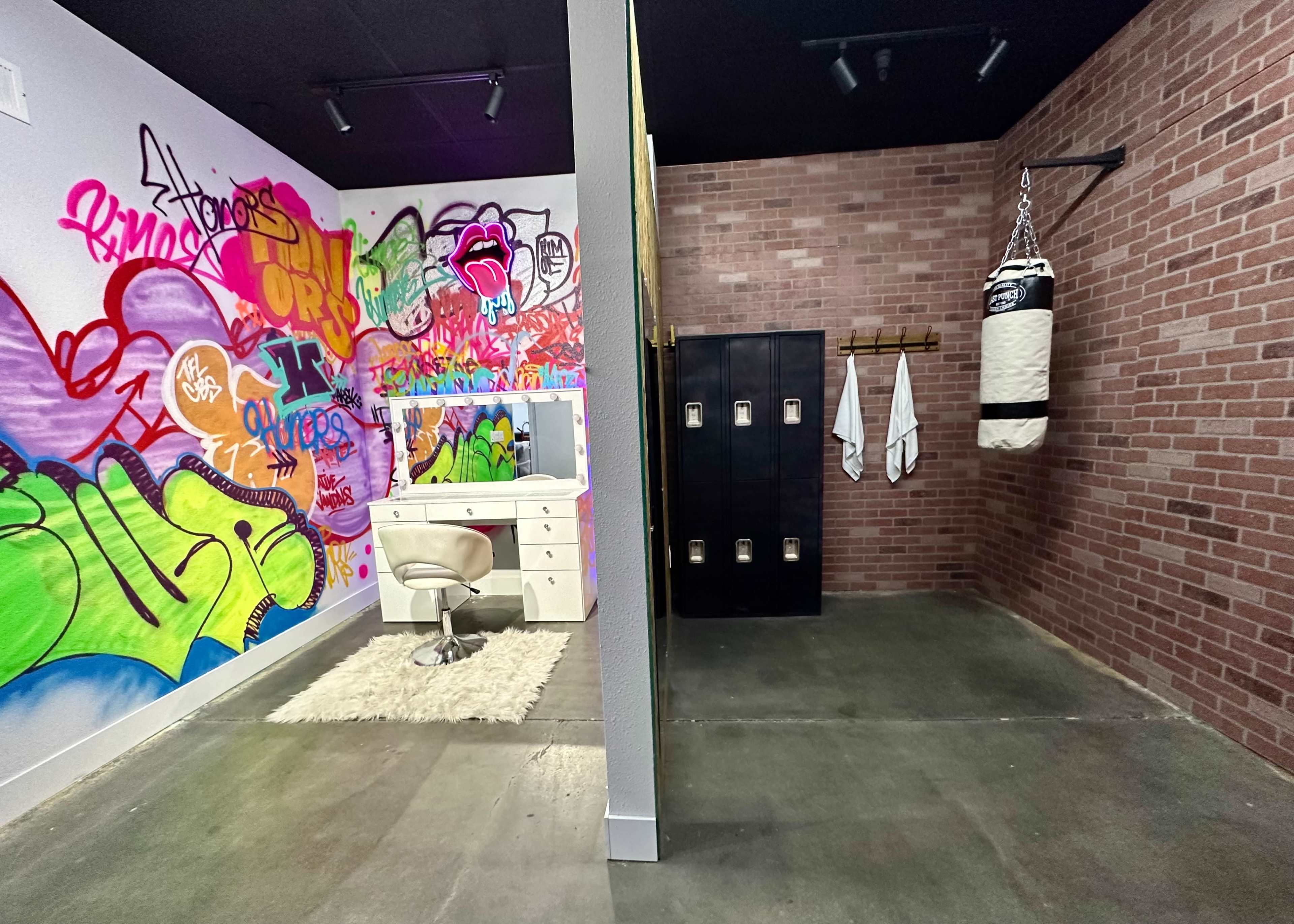 A room divided by a wall, featuring a colorful graffiti mural on one side with a vanity and chair, and a boxing area with lockers and a punching bag on the other side.