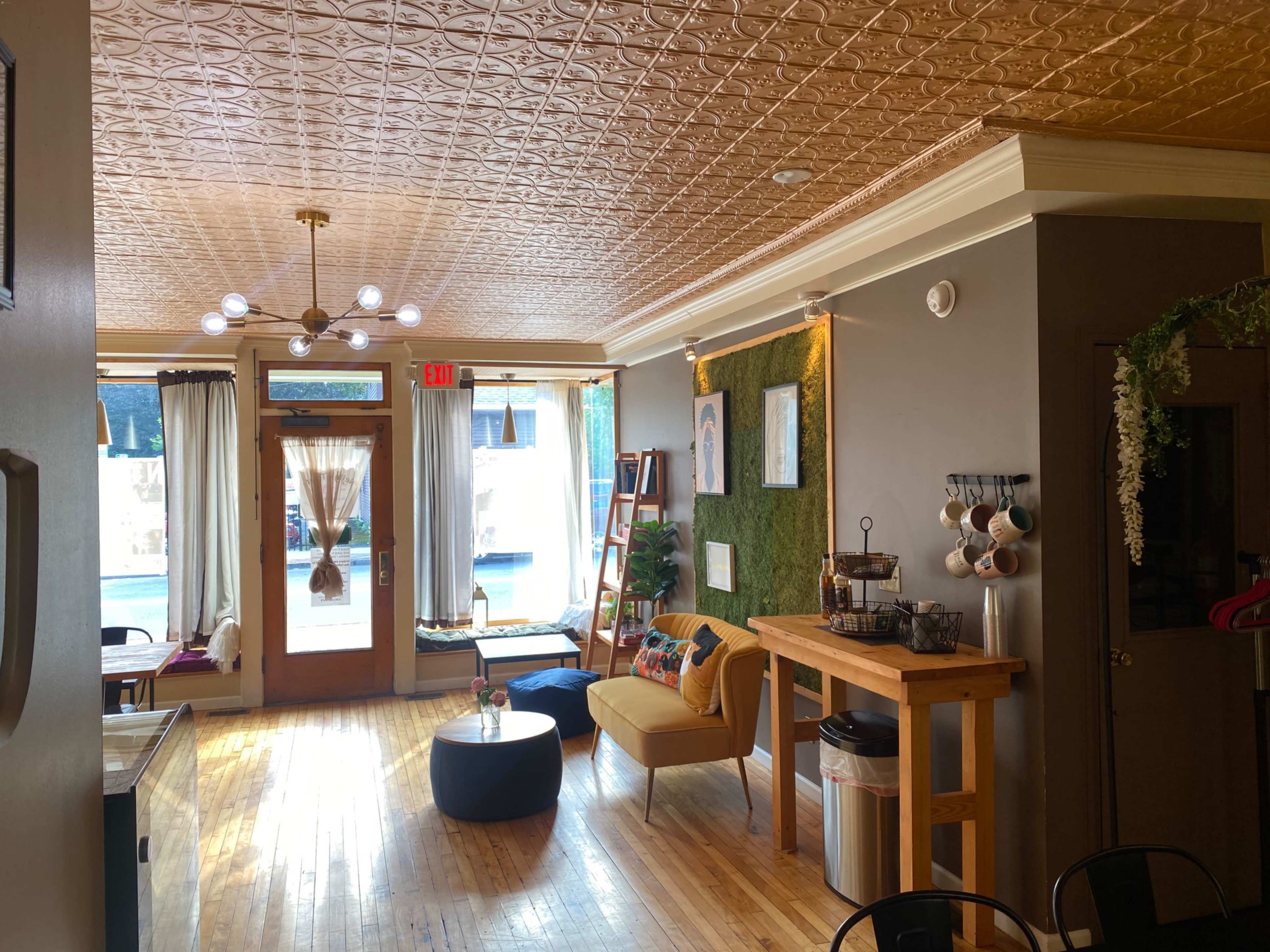 The image shows a cozy interior of a café or common area with wooden flooring, a decorative ceiling, and a mix of seating options including chairs and a table near a large window.