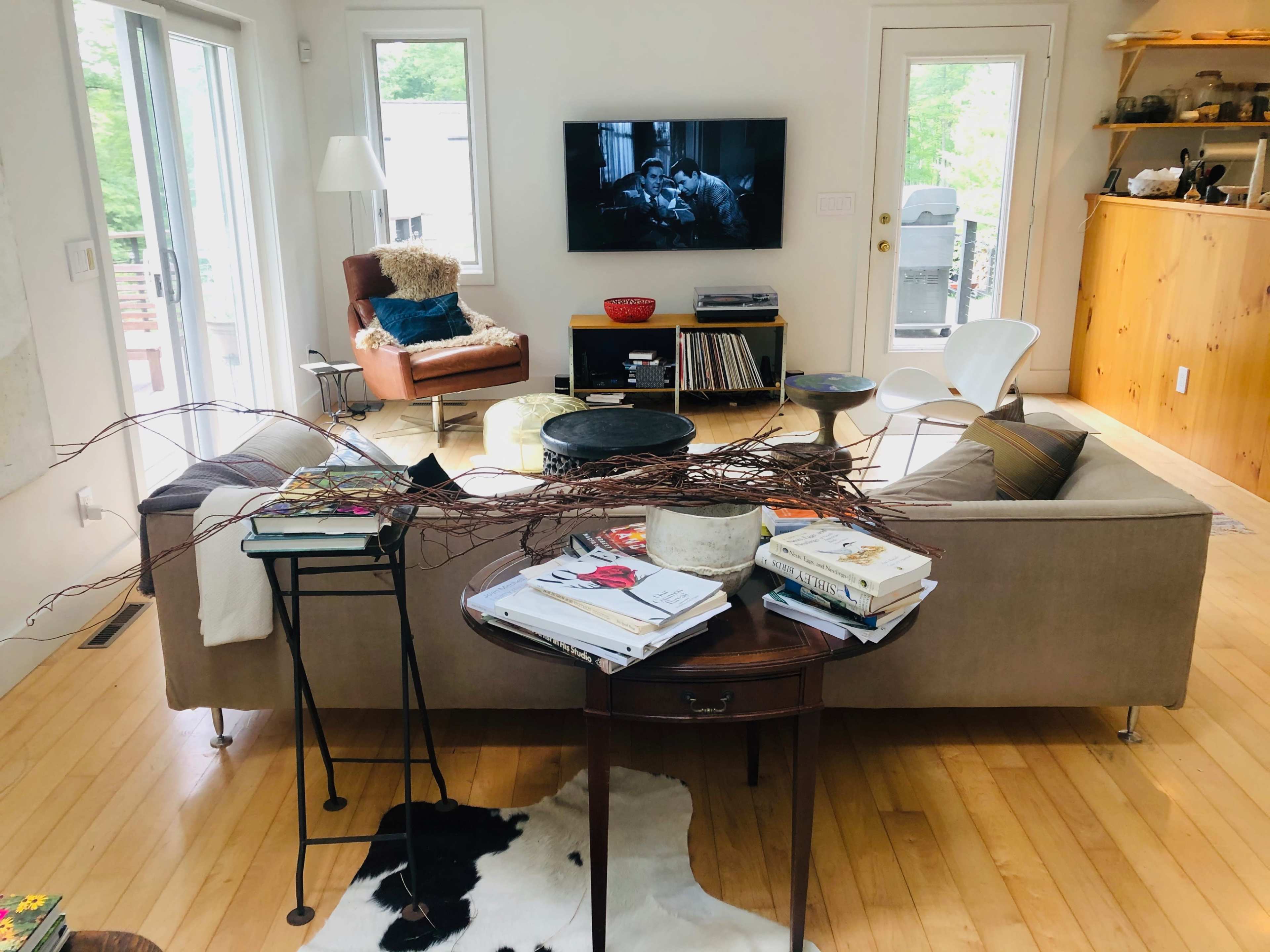 A living room features a beige sofa with a cluttered table, a television displaying a black-and-white film, and a mix of modern and rustic furniture.