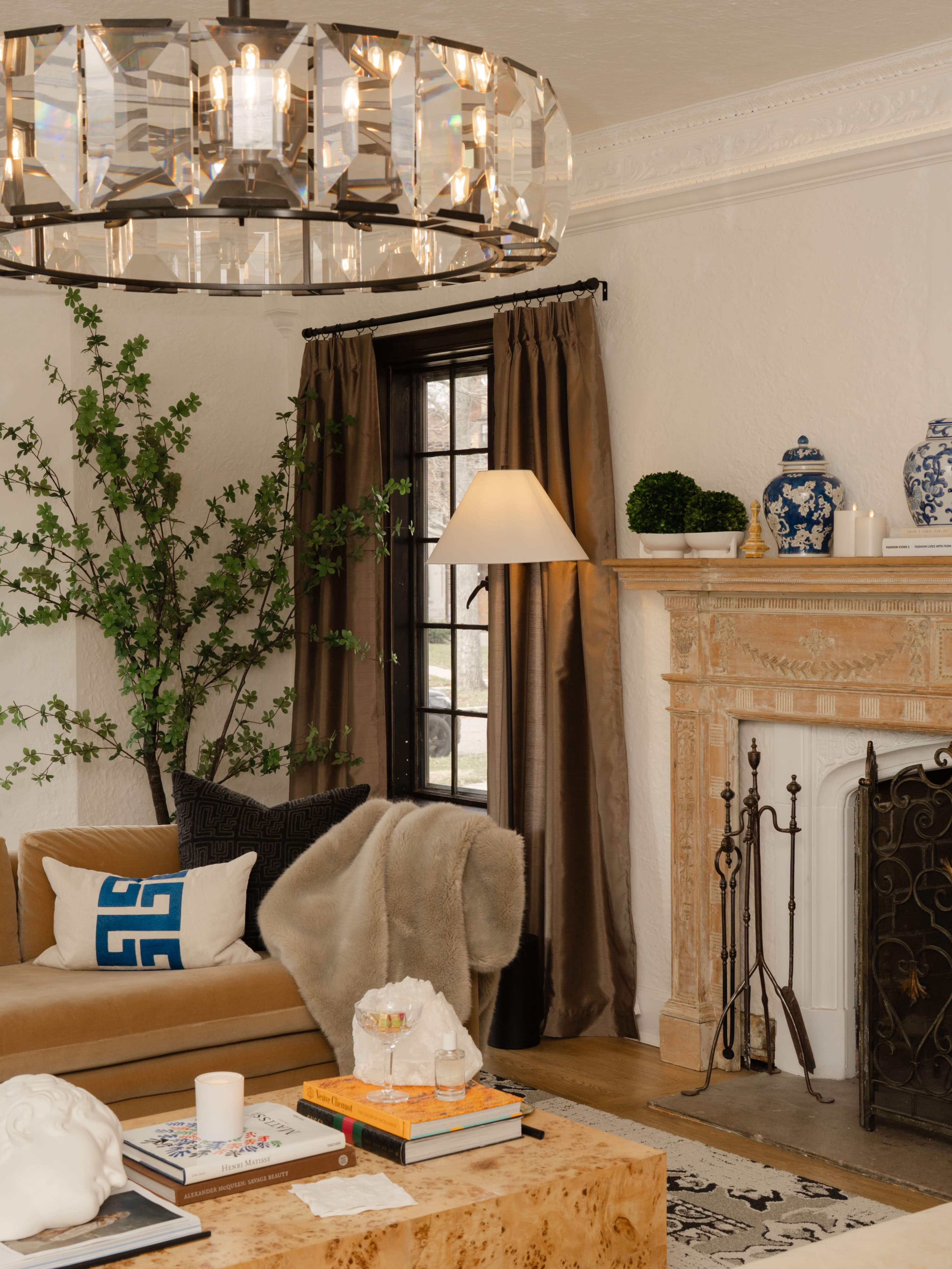 The image shows a stylish living room featuring a large chandelier, a beige sofa with a blue accent pillow, and a traditional fireplace adorned with decorative items and greenery.