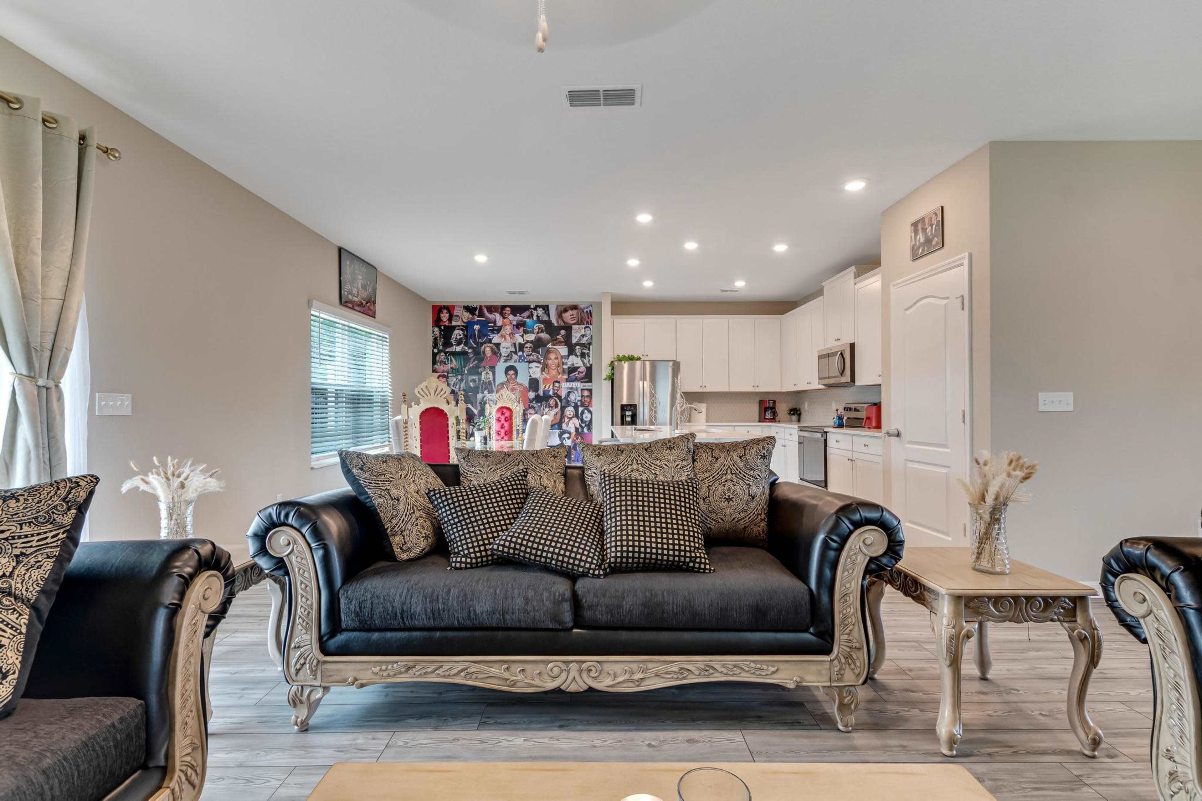A living room featuring a black couch with patterned cushions, a coffee table, and a kitchen area in the background adorned with a collage of photographs.