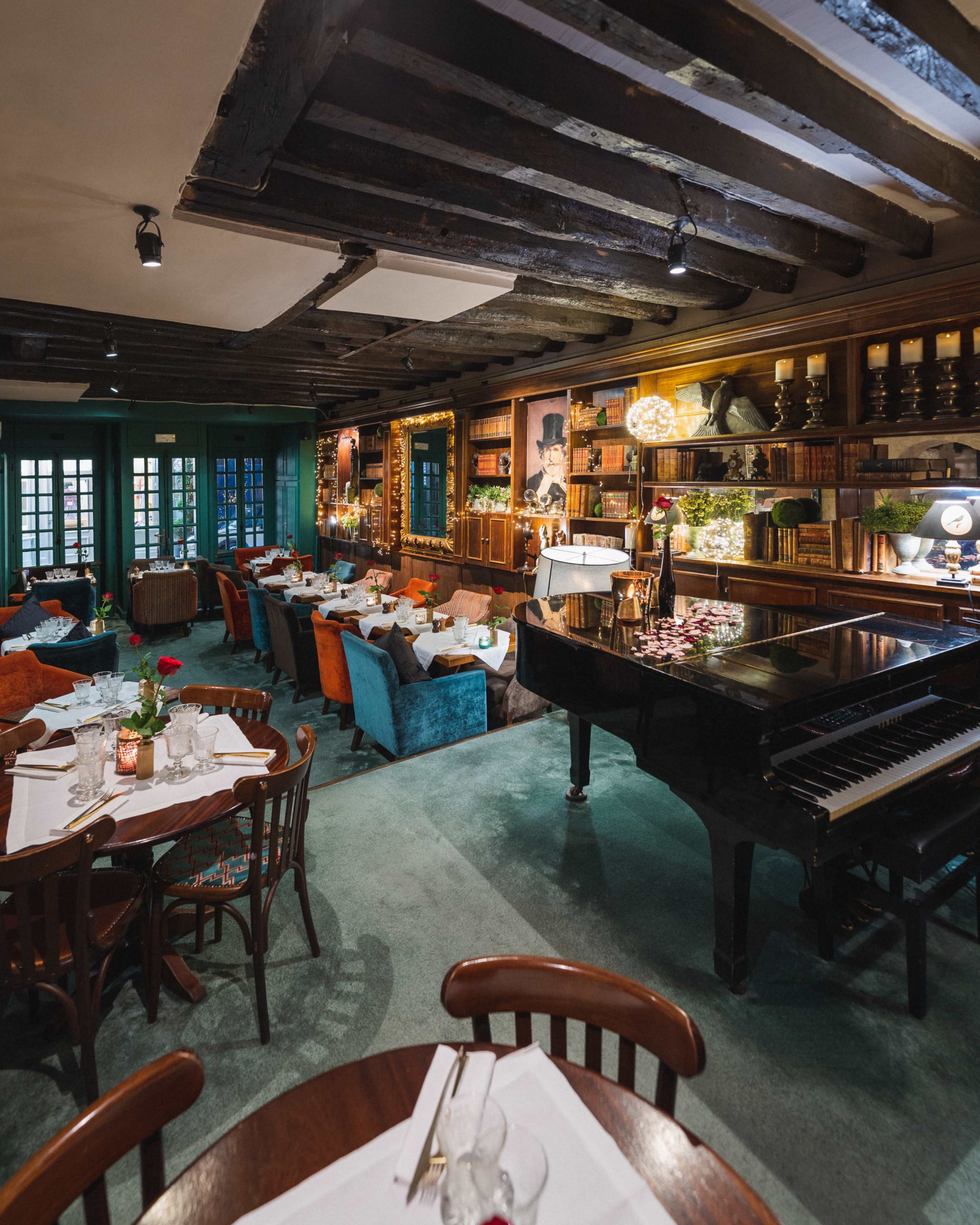 The image shows a cozy restaurant interior featuring dark wooden beams, multiple dining tables, and a grand piano.