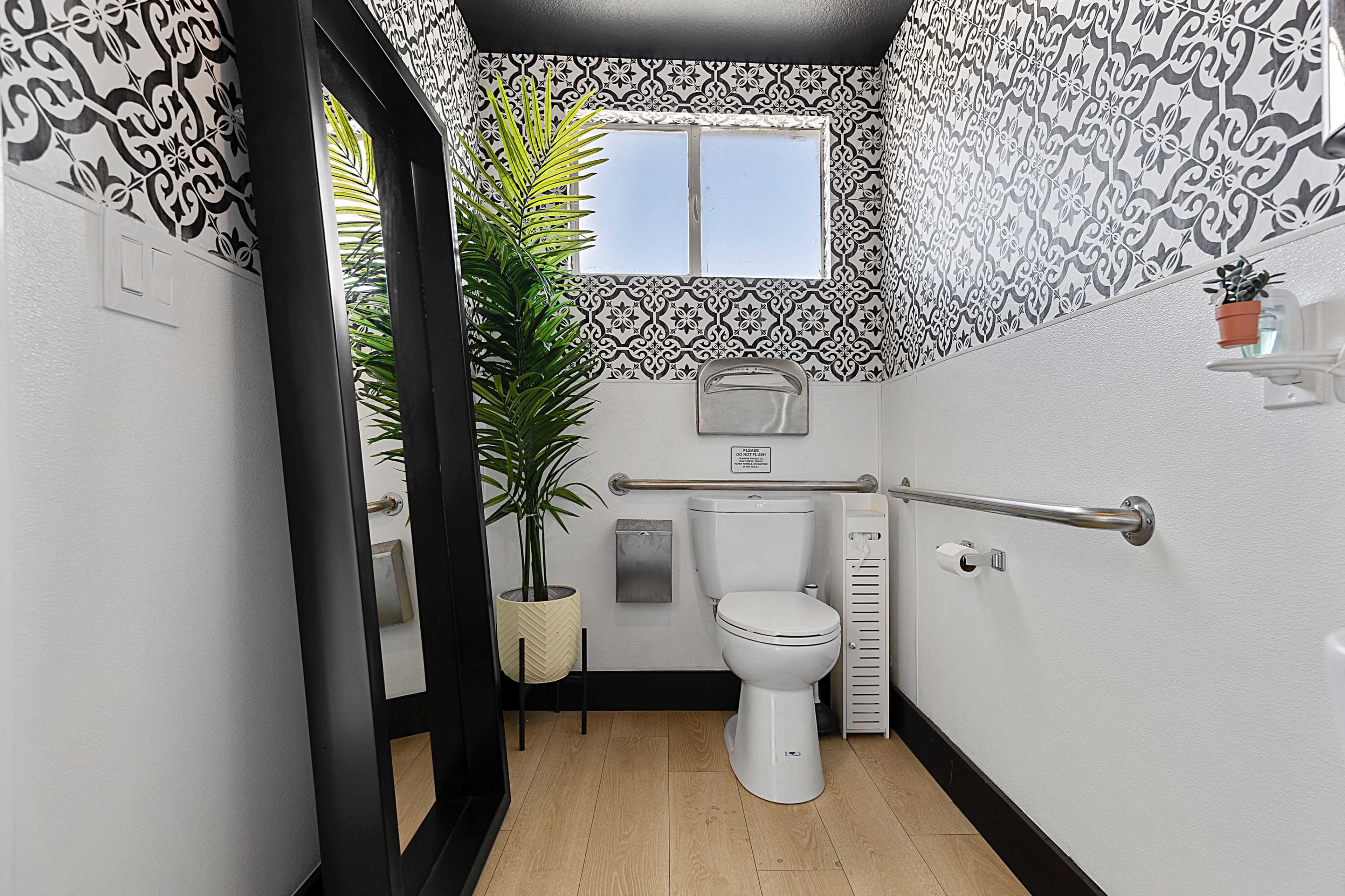 A small bathroom with a toilet, a large mirror, decorative wall patterns, a window, and a potted plant.