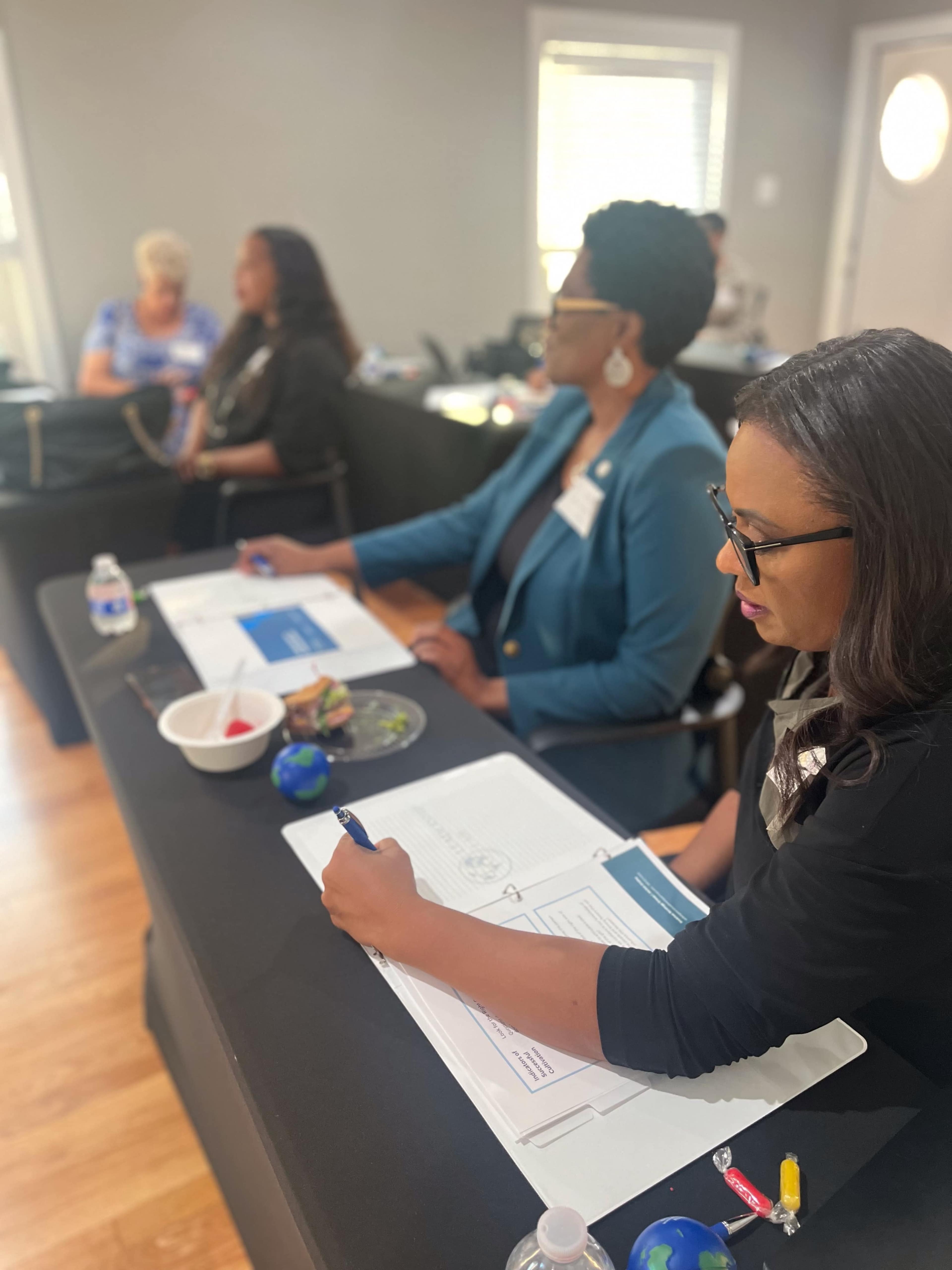 The image shows two women seated at a table with documents and a small dish, while others are engaged in discussion in the background.