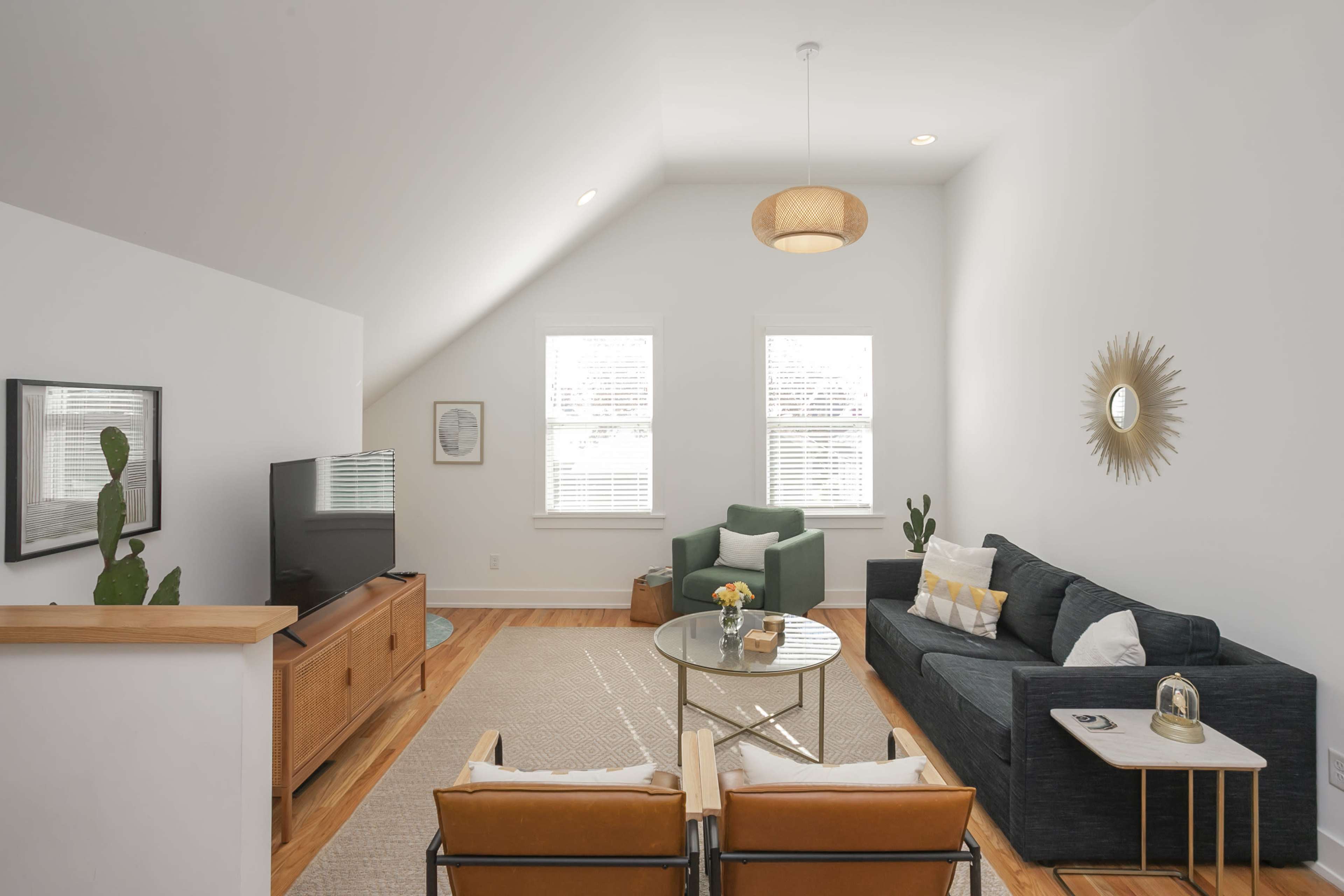 The living room features a modern design with a dark sofa, two orange chairs, a round coffee table, and a television on a wooden console, all set against white walls and natural light from the windows.