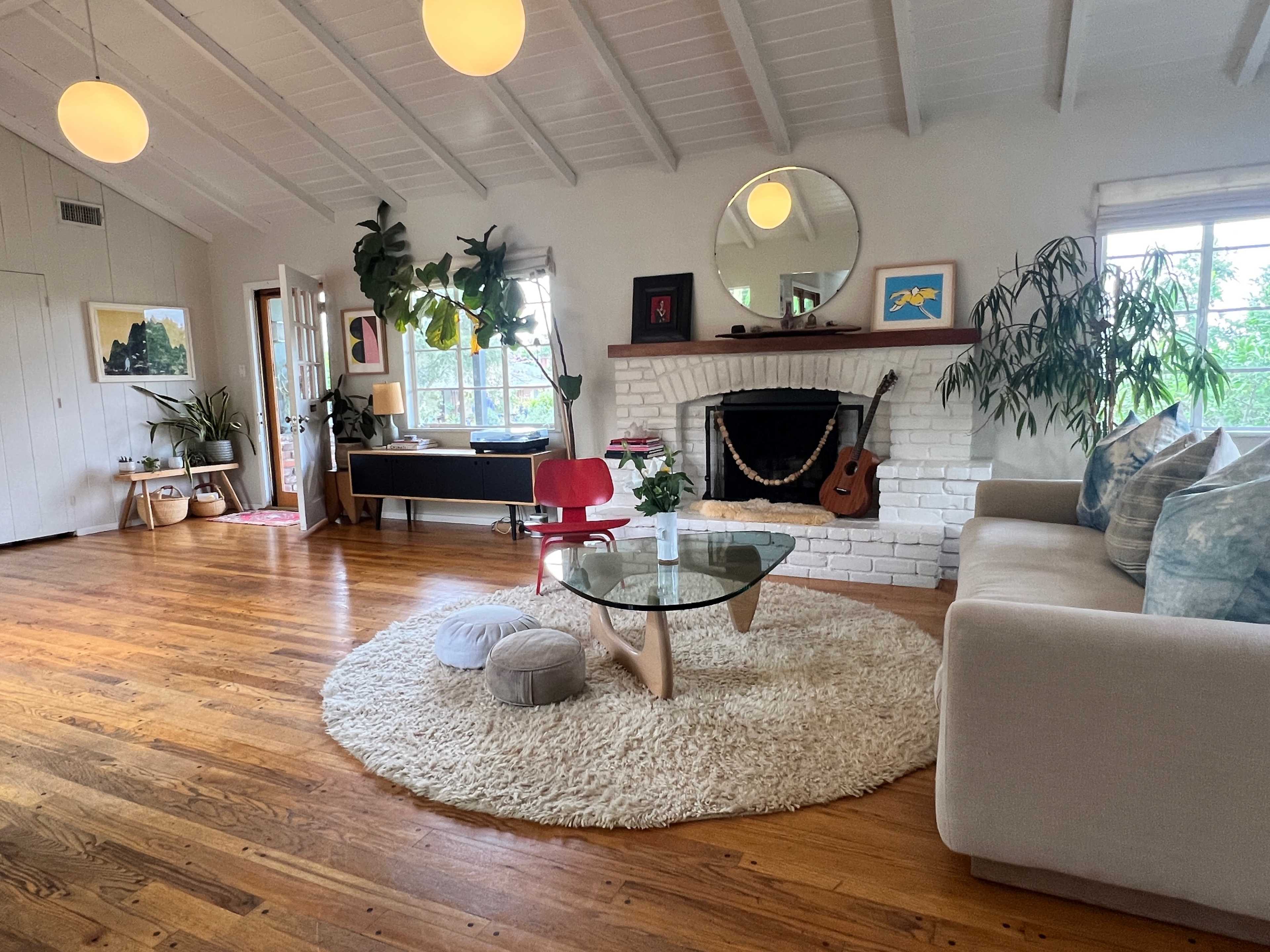 A cozy living room with a white brick fireplace, a circular coffee table on a large rug, and a mix of plants and artwork decorating the space.