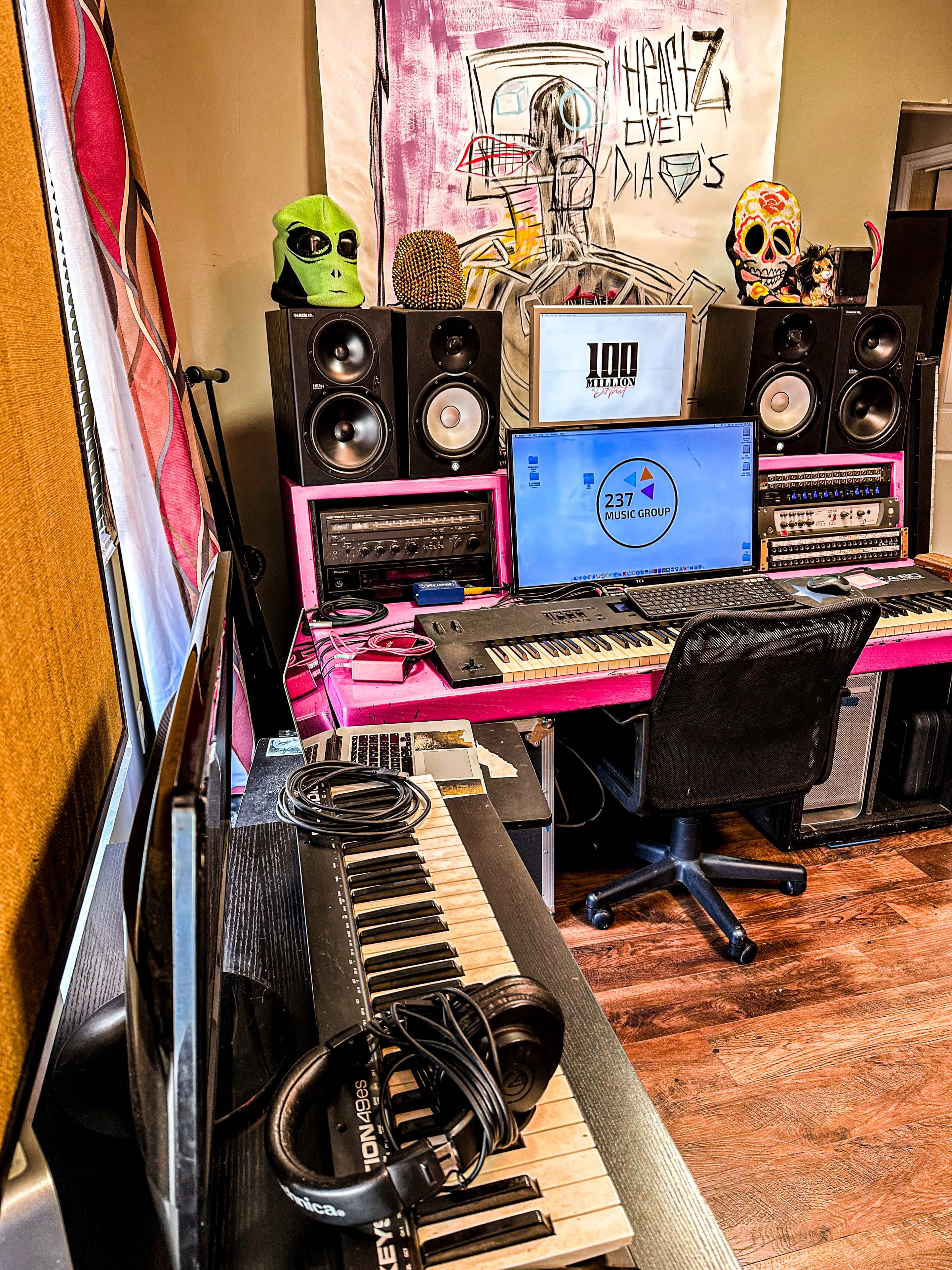 The image shows a music production studio with a computer, keyboards, speakers, and decorative skulls on display.