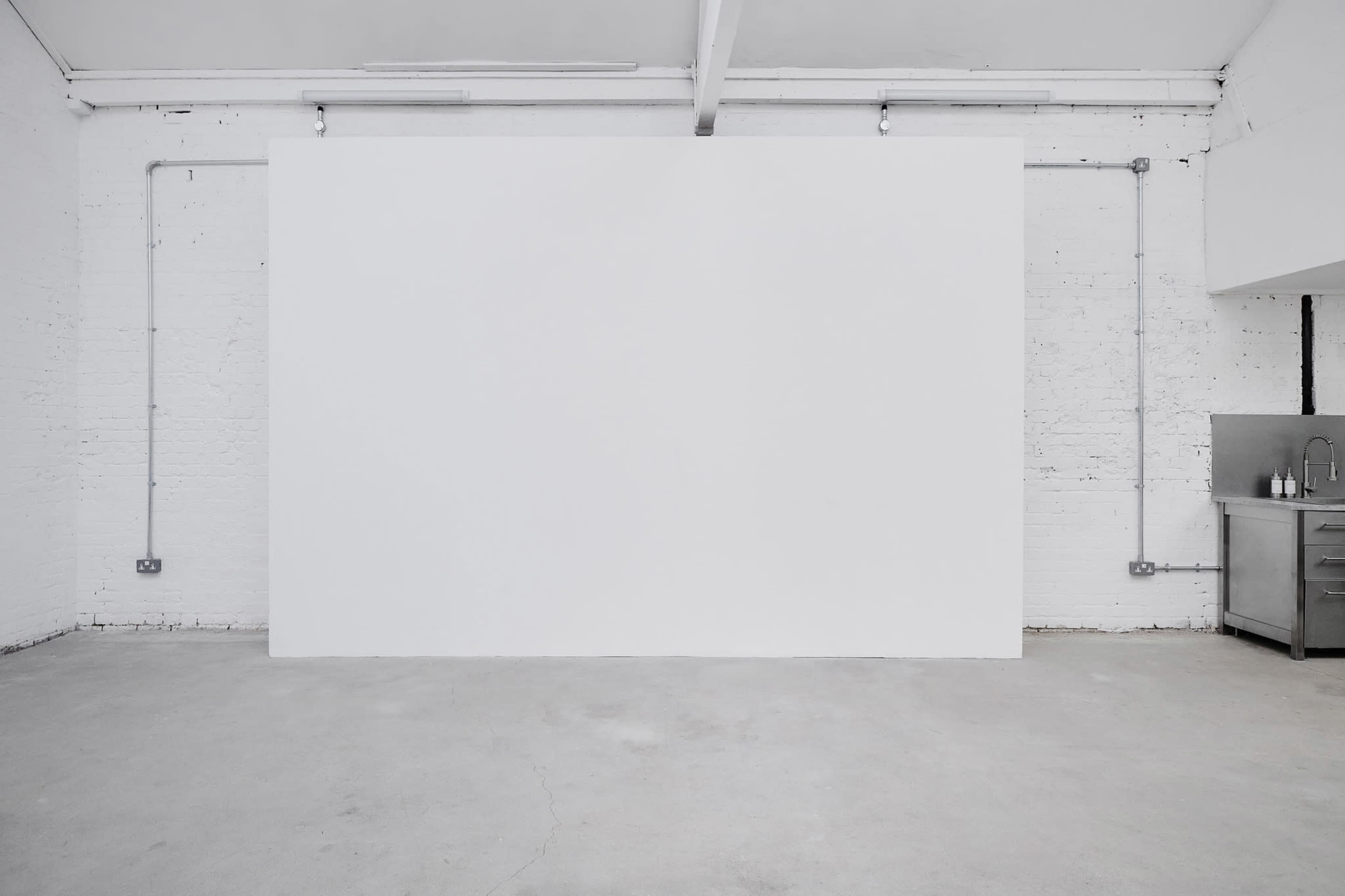 The image shows a spacious, empty room with a large white wall, concrete flooring, and a stainless steel counter in one corner.