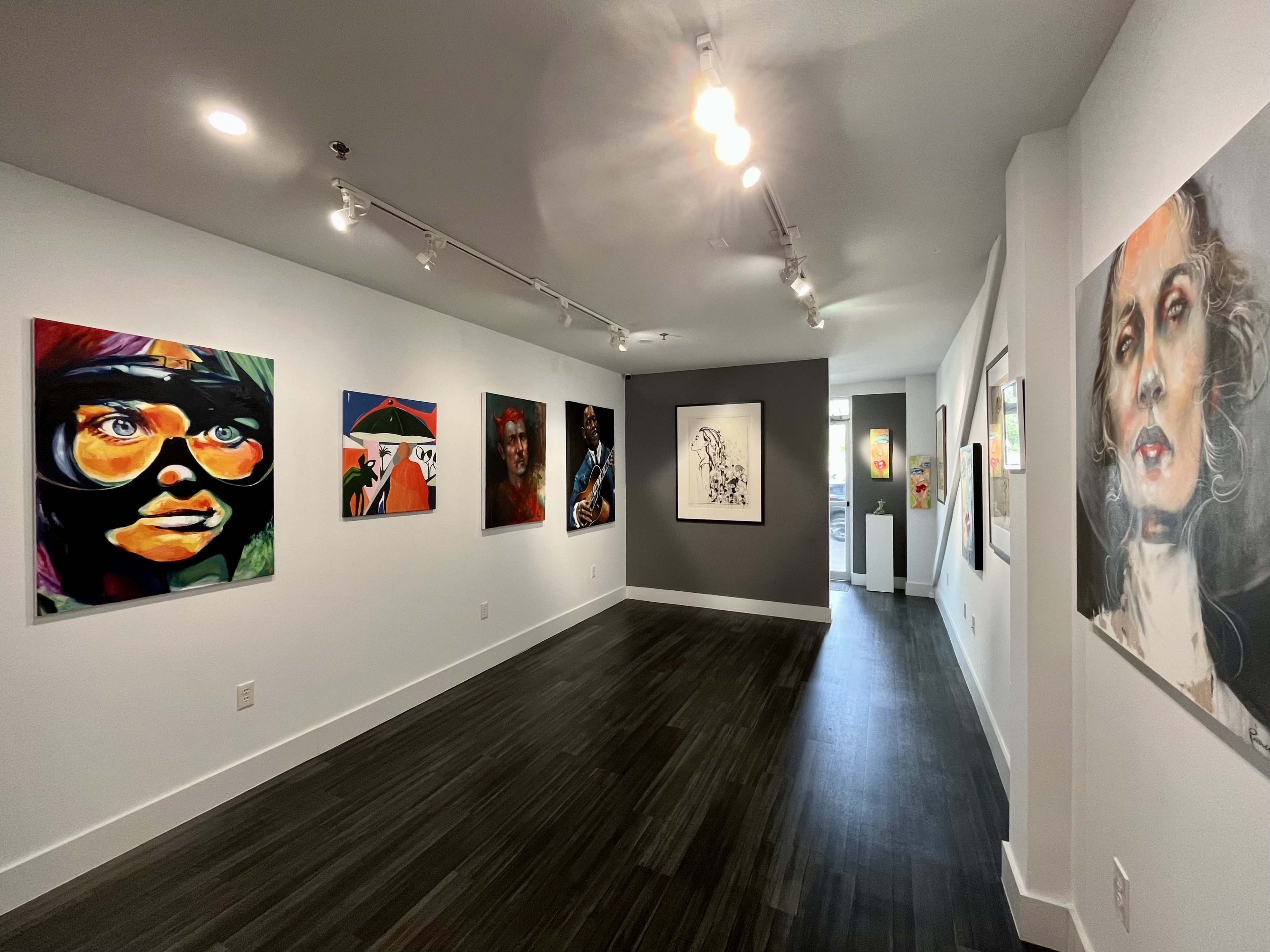 An art gallery interior featuring several colorful paintings on the walls with a corridor leading to another room at the end.