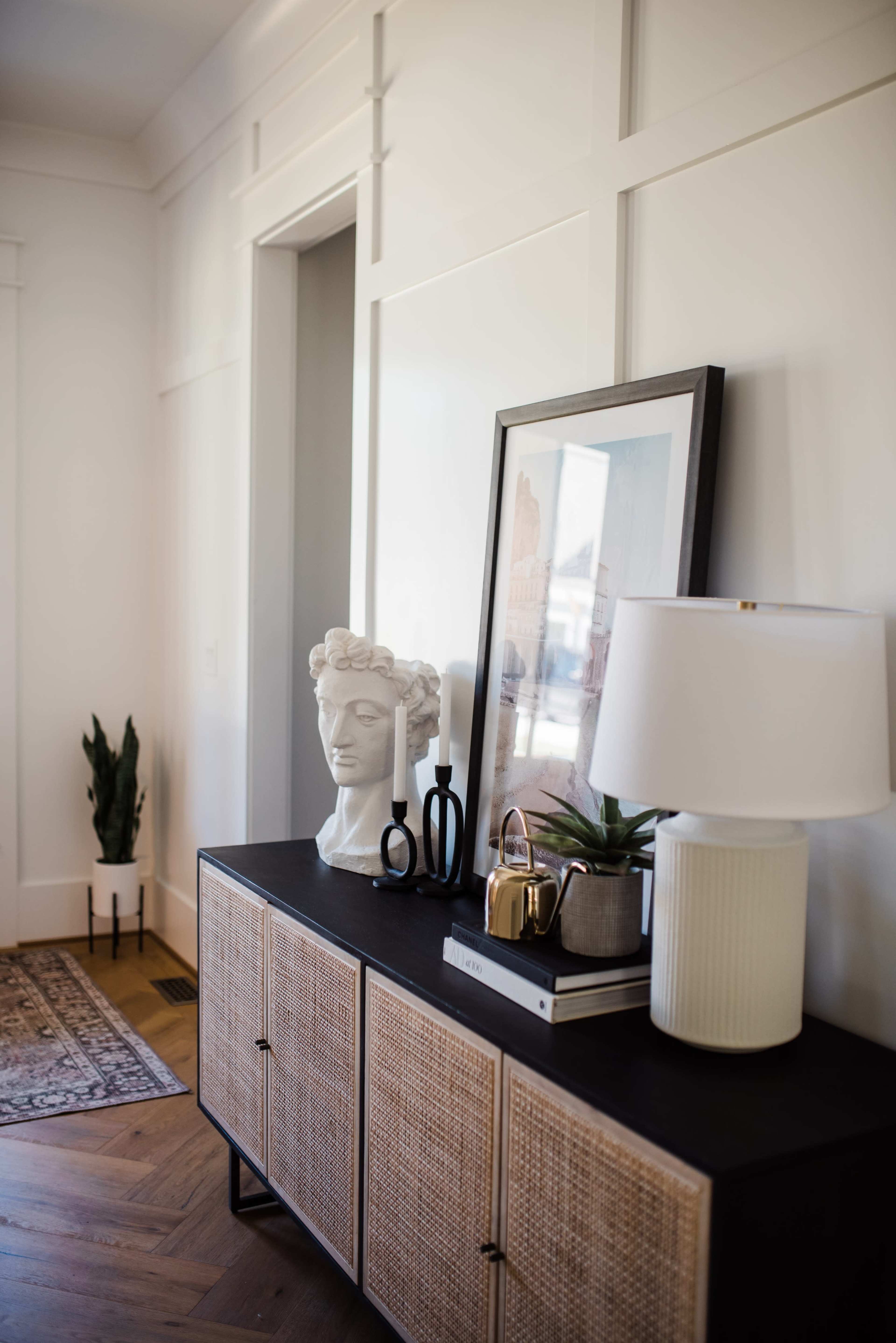 A console table with a sculpture, framed artwork, a plant, and a lamp is positioned against a white paneled wall in a well-lit room.