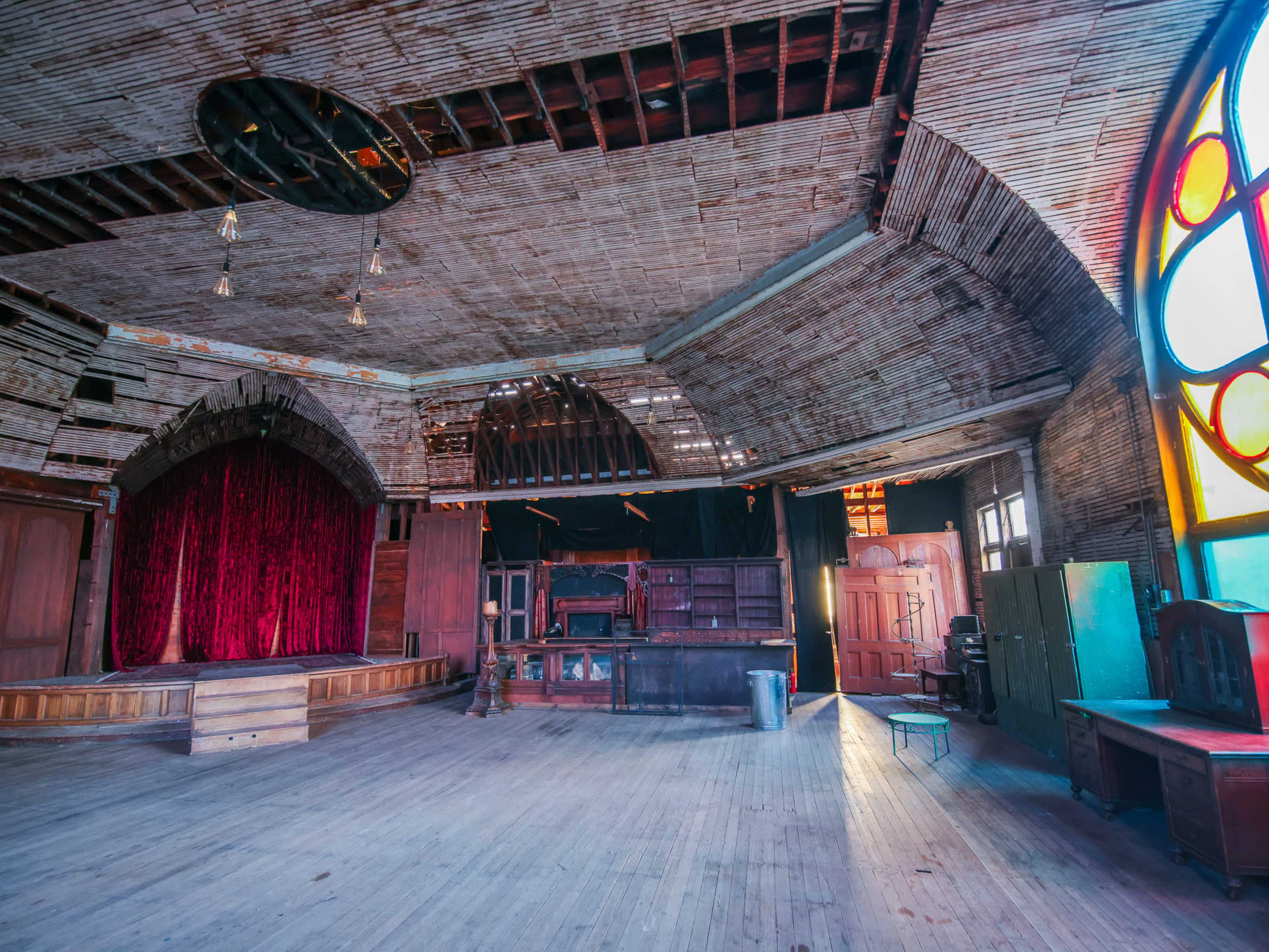 The image shows an old, empty theater with a stage, red curtains, and large stained glass windows.