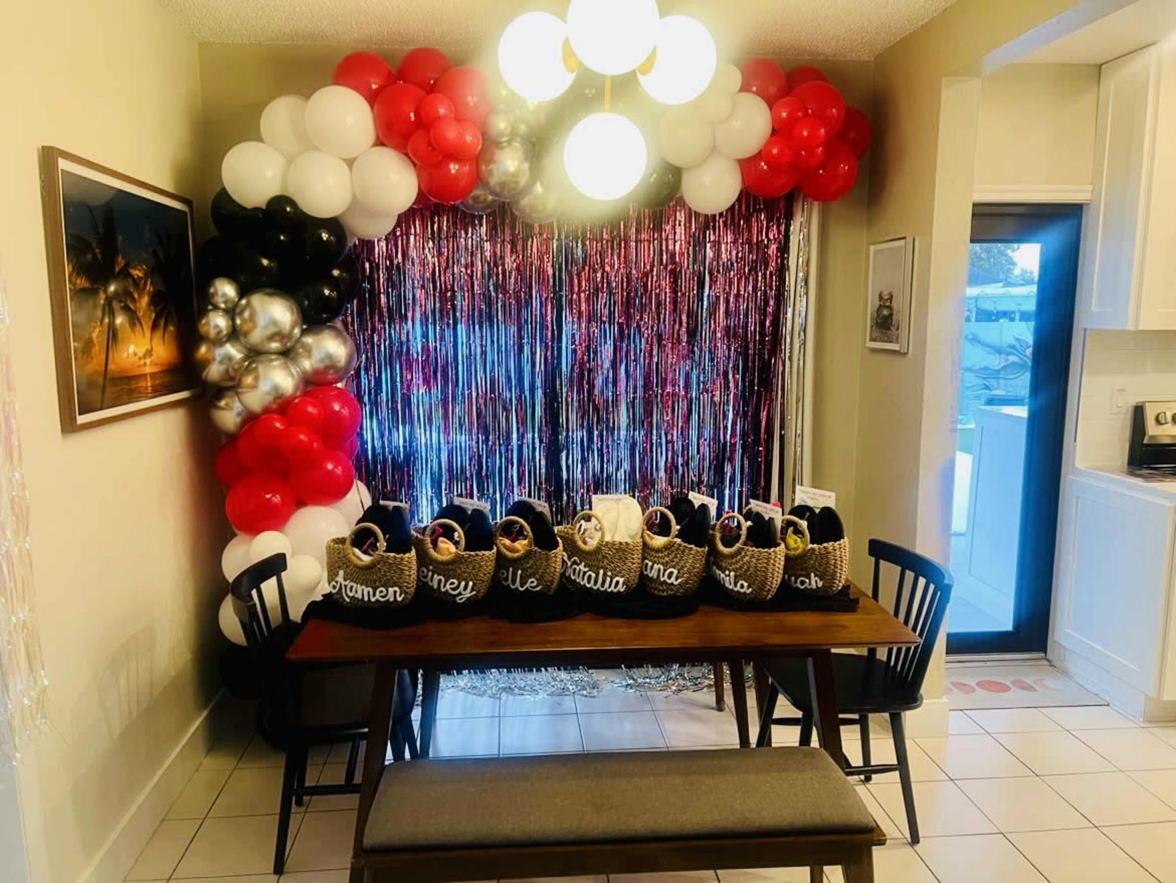 The image shows a decorated dining area with a table set for a celebration, featuring a backdrop of shiny streamers and a colorful balloon arch above it.