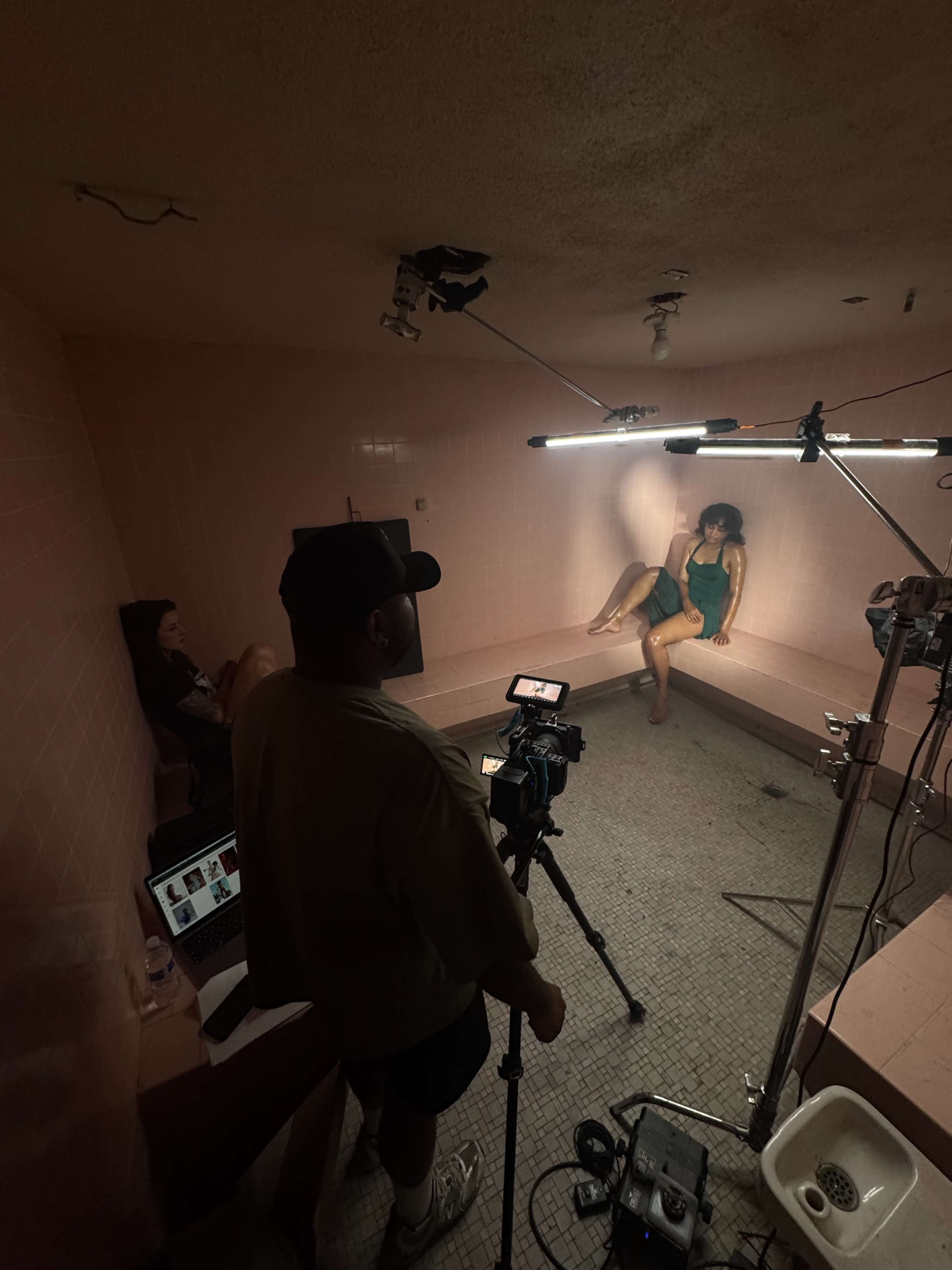 A photoshoot is taking place in a pink-tiled room with two women, one posing and the other sitting nearby, while a photographer sets up equipment.