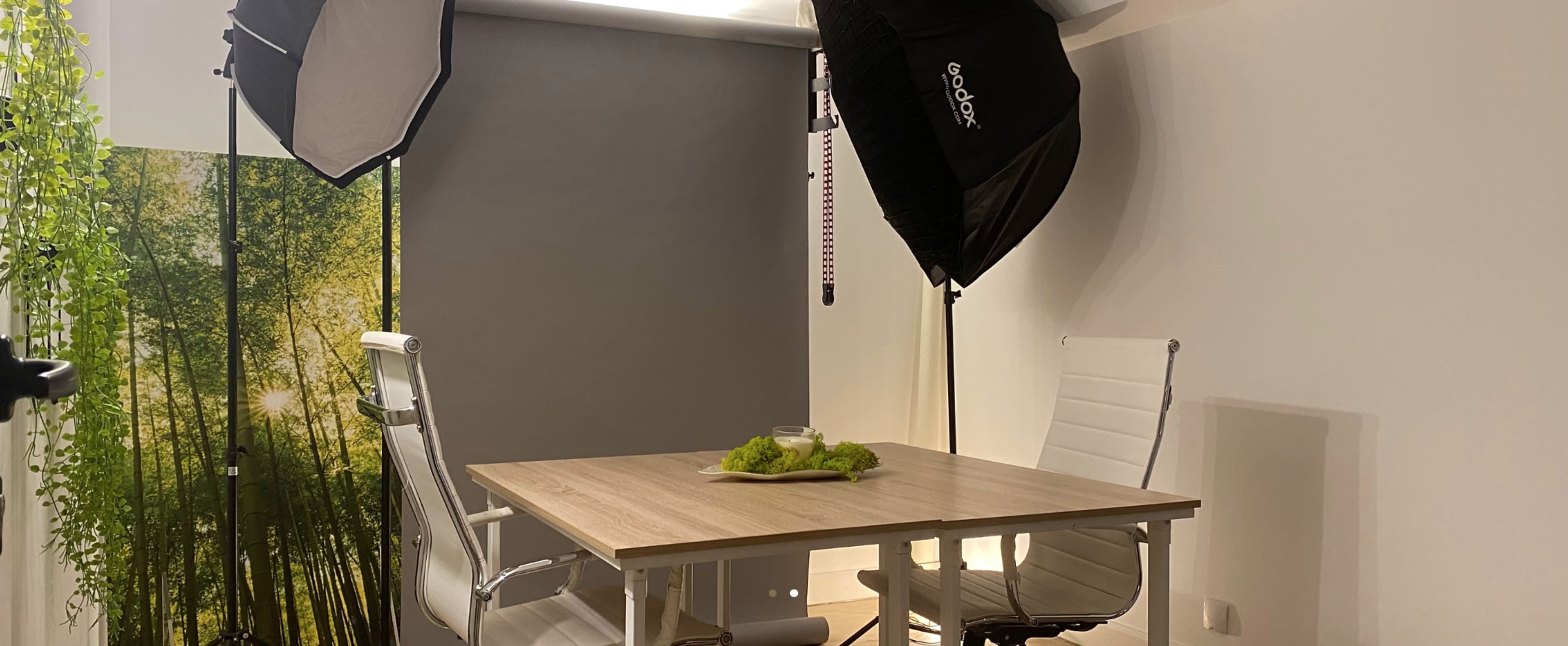 A small photography studio with a table surrounded by white chairs, complemented by soft lighting and a backdrop featuring greenery.