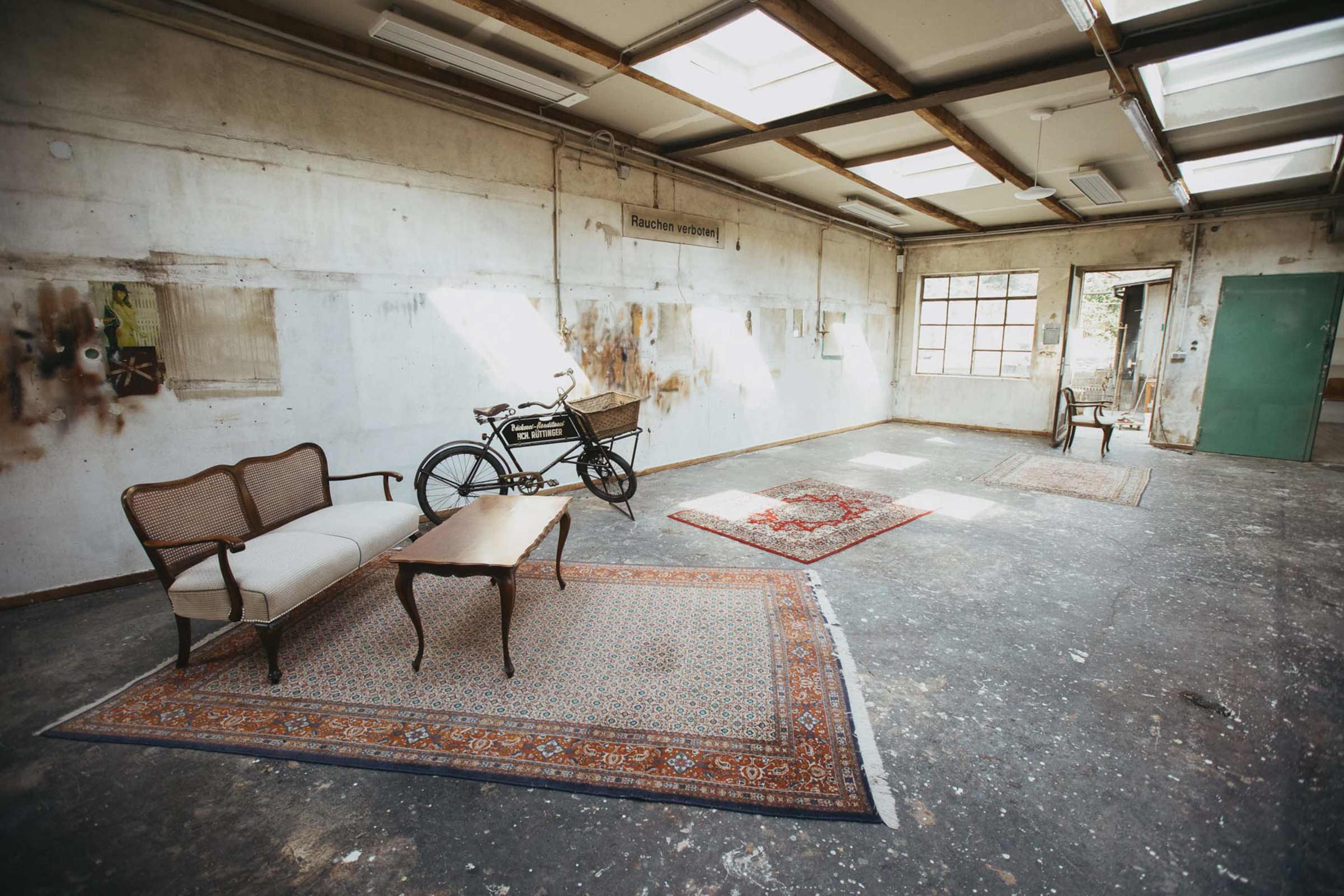 The image shows an empty, industrial room with a wooden ceiling and skylights, featuring a bicycle, a table, and two armchairs arranged on patterned rugs.