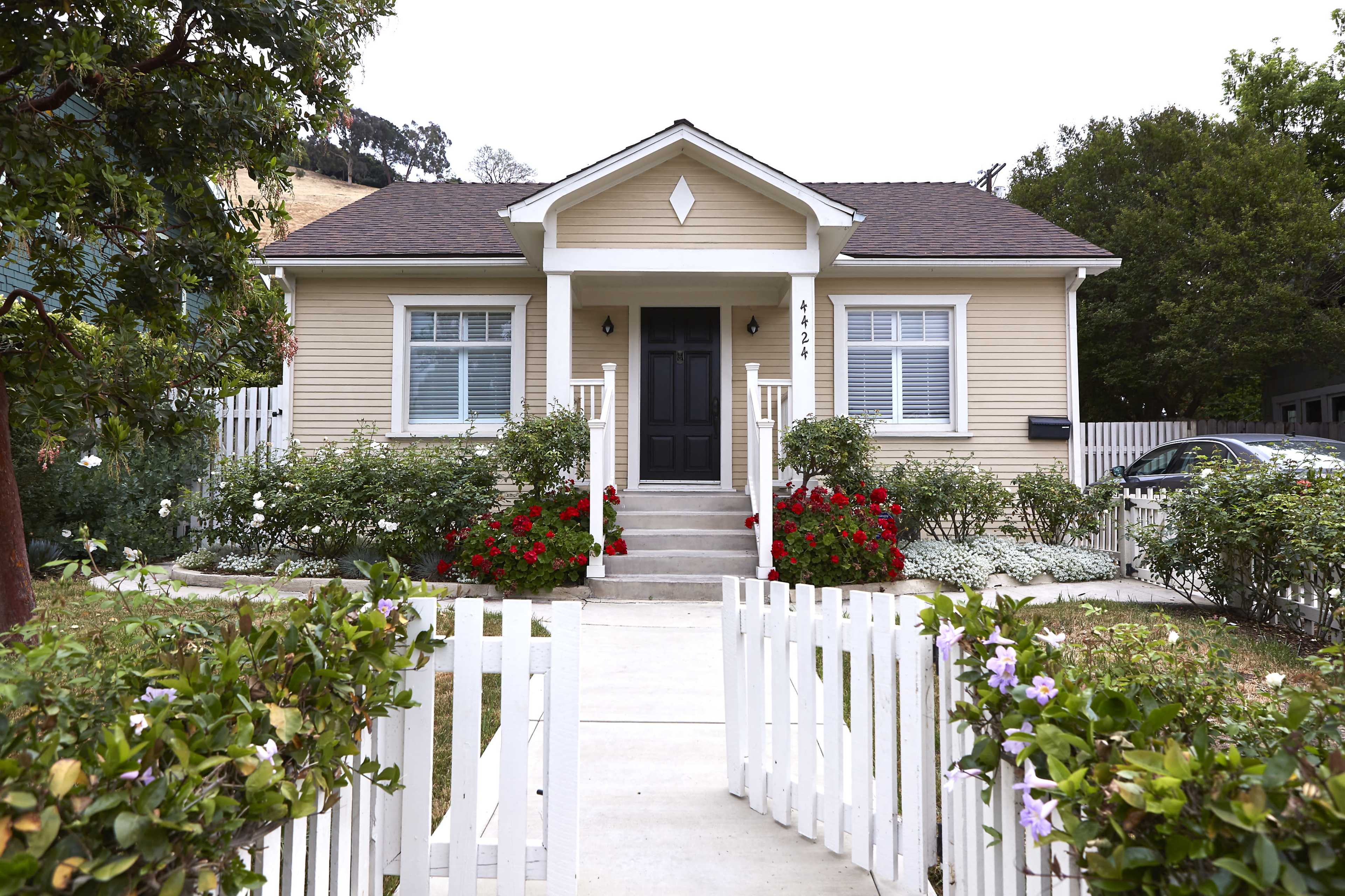 A small, light yellow house with a black front door is surrounded by a white picket fence and colorful flower beds.