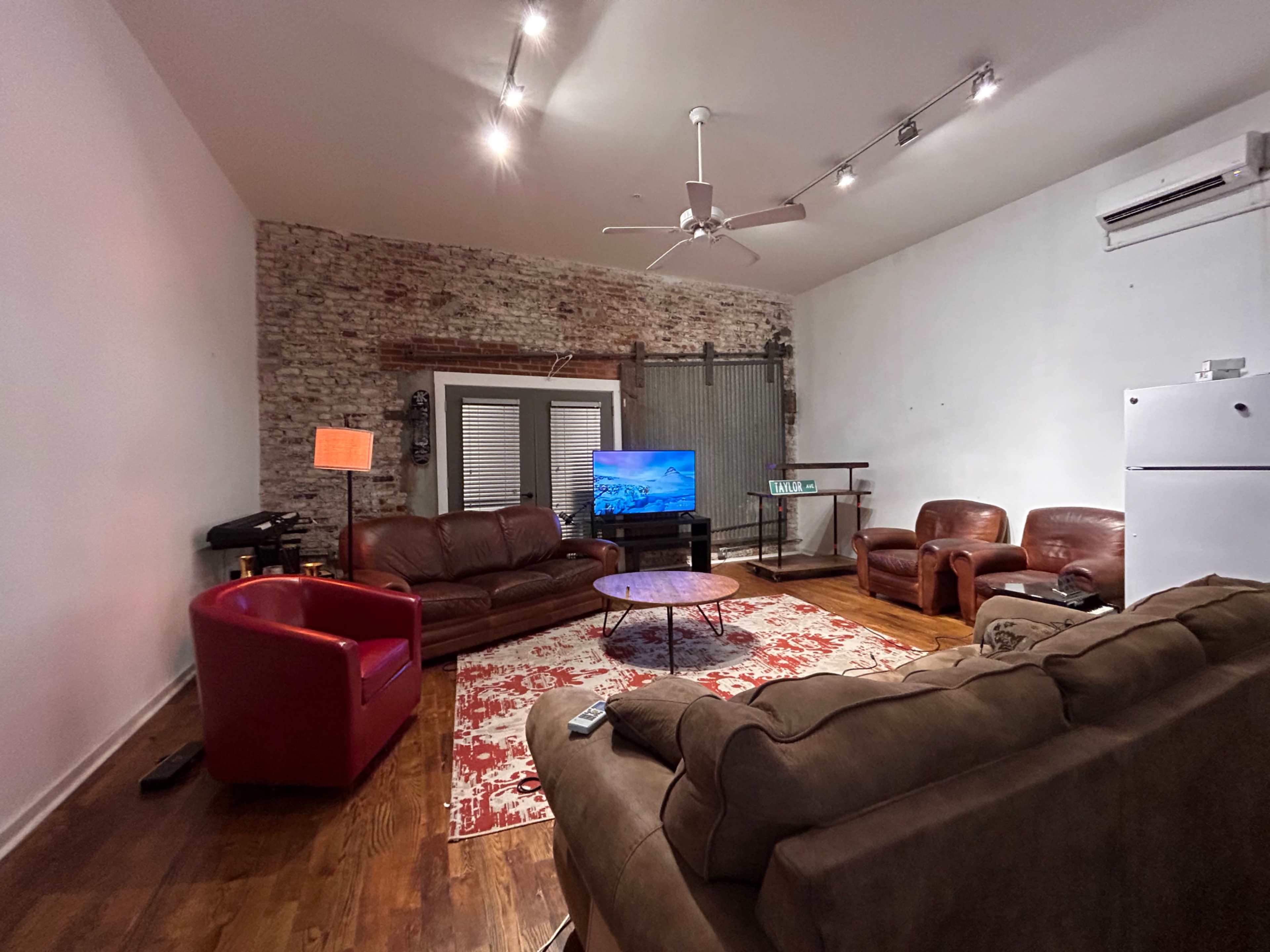 A living room with a large area rug, multiple seating options, a television on a stand, and exposed brick walls.