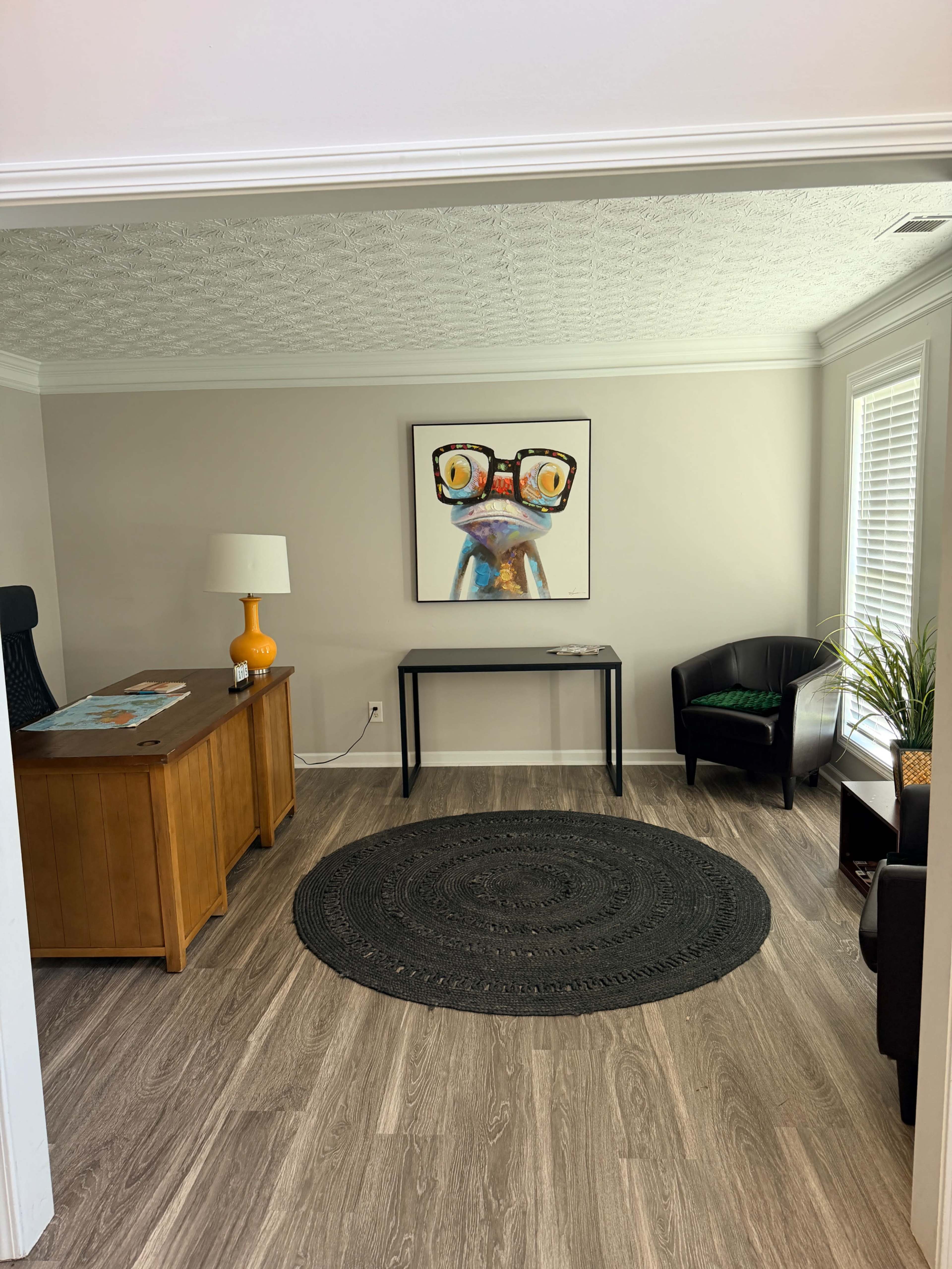 The image shows a small office space featuring a wooden desk, a black round rug, a chair, and a colorful painting of a frog wearing glasses on the wall.