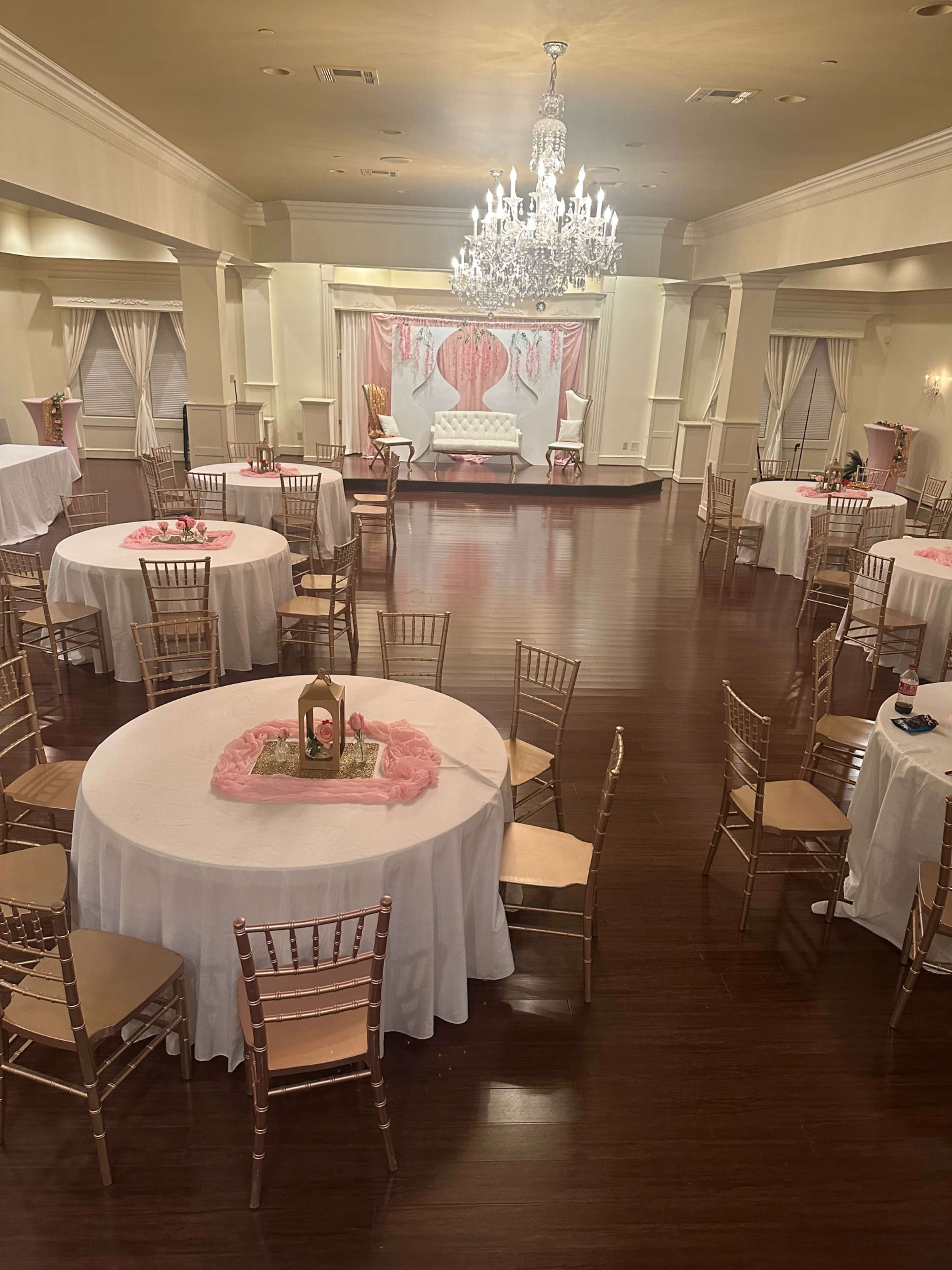 A spacious event hall is set up with several round tables, each adorned with white tablecloths and decorative centerpieces, while the stage at the front features a light-colored backdrop and a crystal chandelier above.