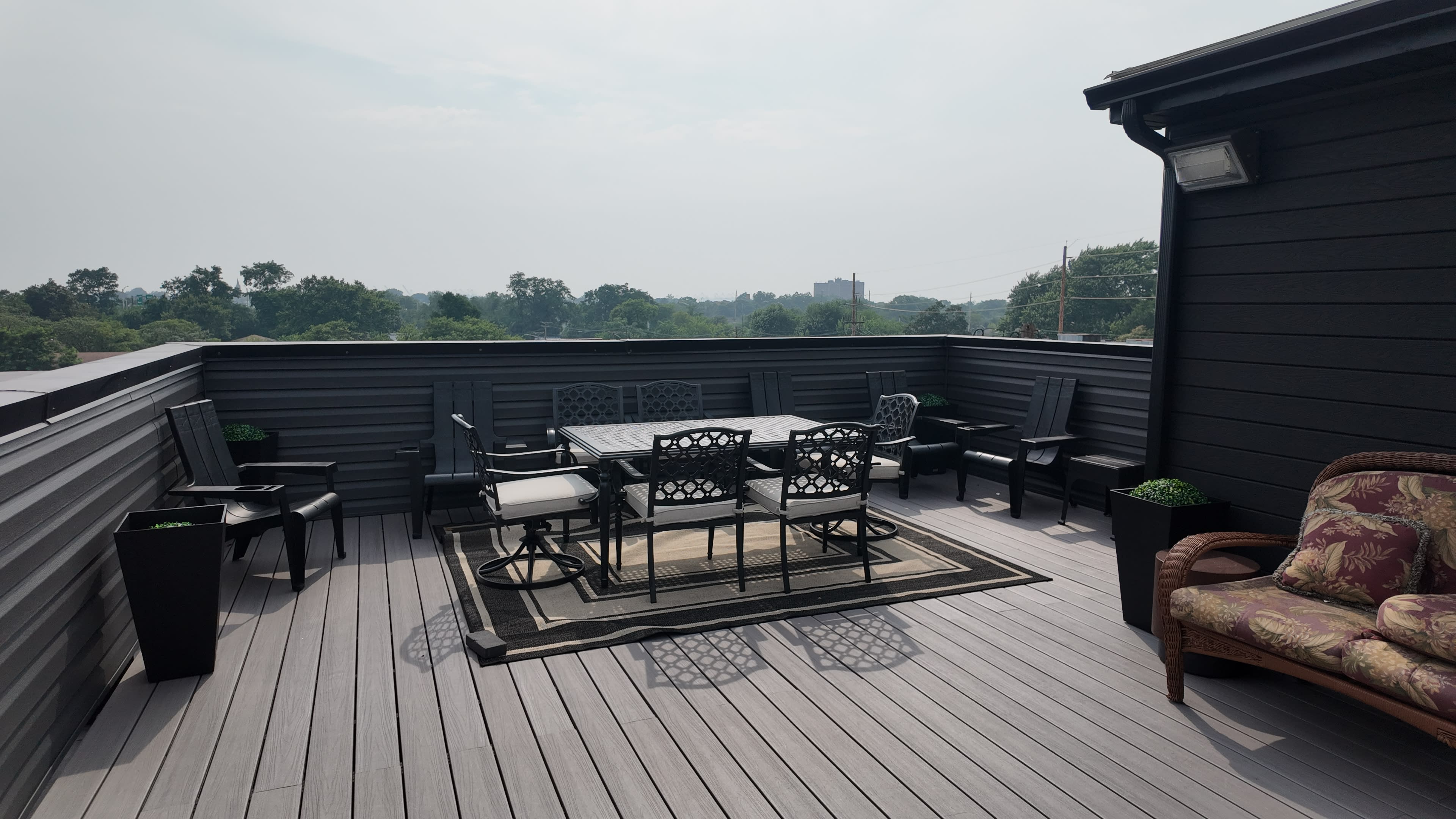 The image shows a rooftop deck with a dining set and seating area surrounded by greenery in the background.